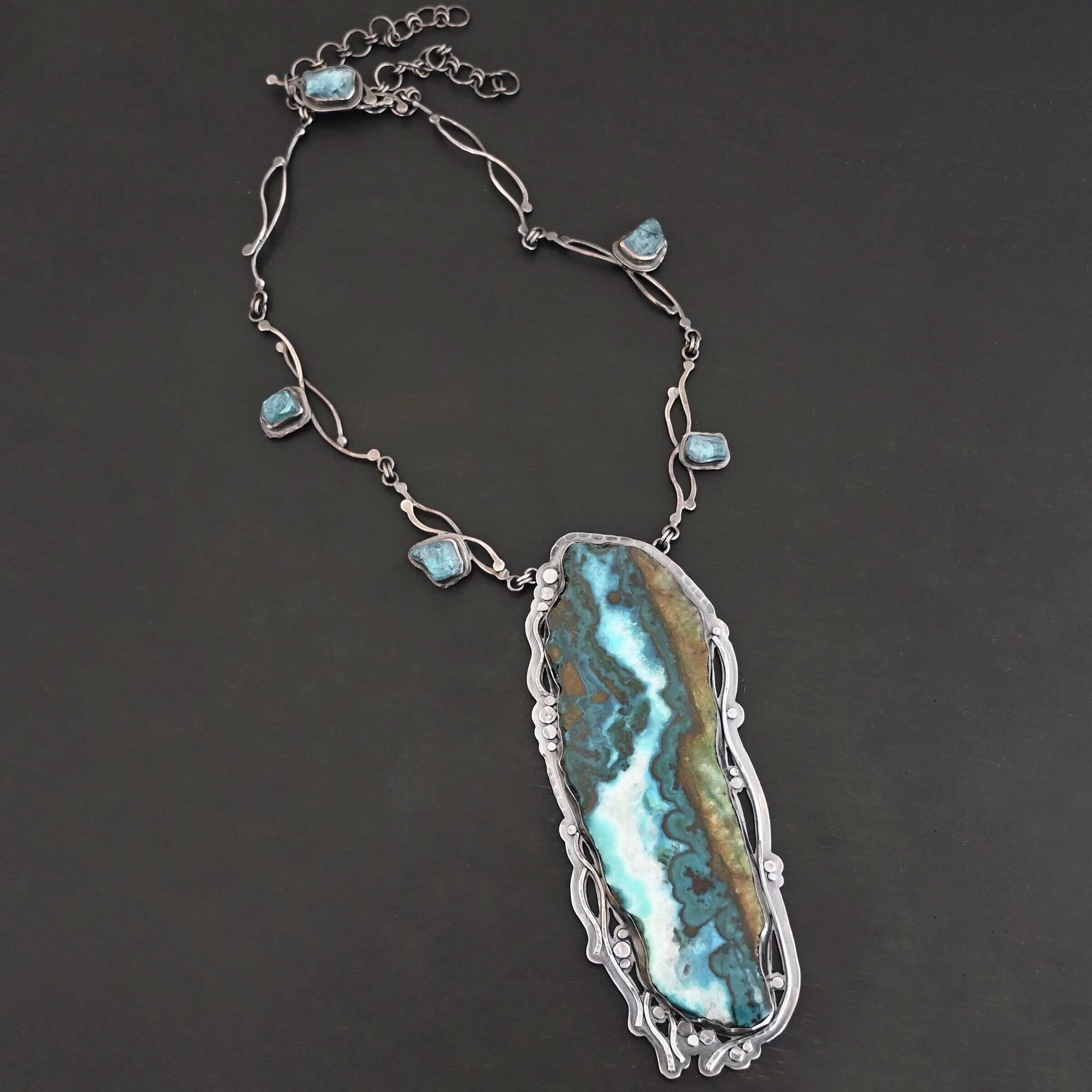 Like The River Runs Necklace Close up of the "Like The River Runs" Necklace. Made out of sterling silver, azurite, and malachite and apatite. The azurite and malachite are mixed within the middle stone. It is a mix of a dark teal, light blue, white, dark green, and light green colors. The light blue and white flow throughout the middle of the stone, and the dark and light green outline the edges of the stone, so it looks as if there is water flowing through the necklace. It is set in sterling silver. Around the setting are silver dots that are hammered so that they are flat. There are also thin silver lines on the bottom of the necklace that look as if they are flowing downwards. There are two light blue stones on the collar of the necklace, one on the left side and one on the right side. Necklace is set against a dark background.