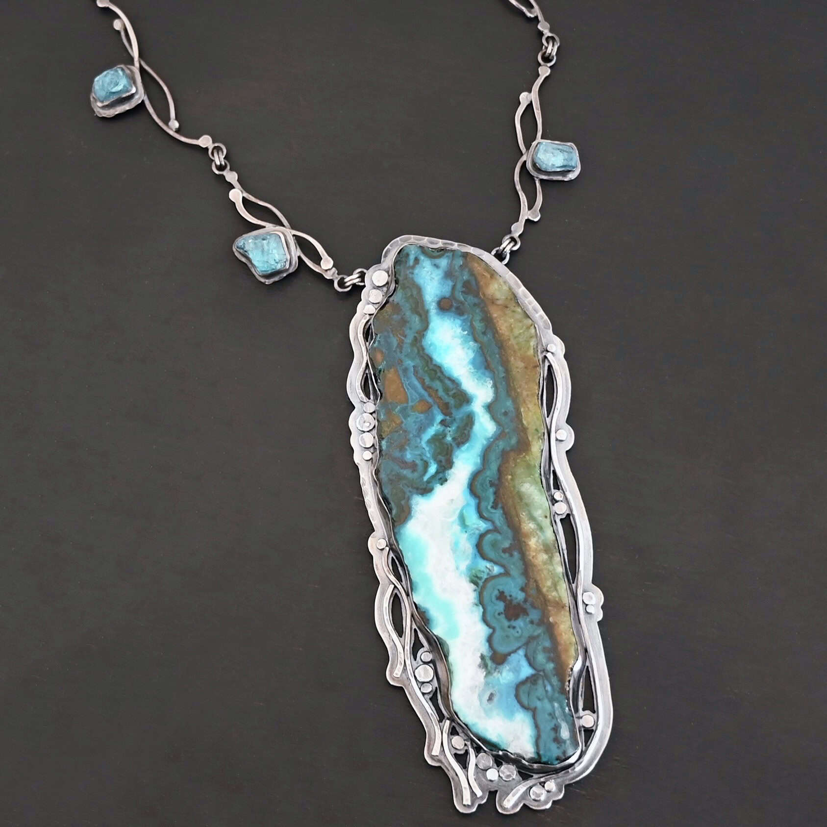 Like The River Runs Necklace Close up of the "Like The River Runs" Necklace. Made out of sterling silver, azurite, and malachite and apatite. The azurite and malachite are mixed within the middle stone. It is a mix of a dark teal, light blue, white, dark green, and light green colors. The light blue and white flow throughout the middle of the stone, and the dark and light green outline the edges of the stone, so it looks as if there is water flowing through the necklace. It is set in sterling silver. Around the setting are silver dots that are hammered so that they are flat. There are also thin silver lines on the bottom of the necklace that look as if they are flowing downwards. There are two light blue stones on the collar of the necklace, one on the left side and one on the right side. Necklace is set against a dark background.