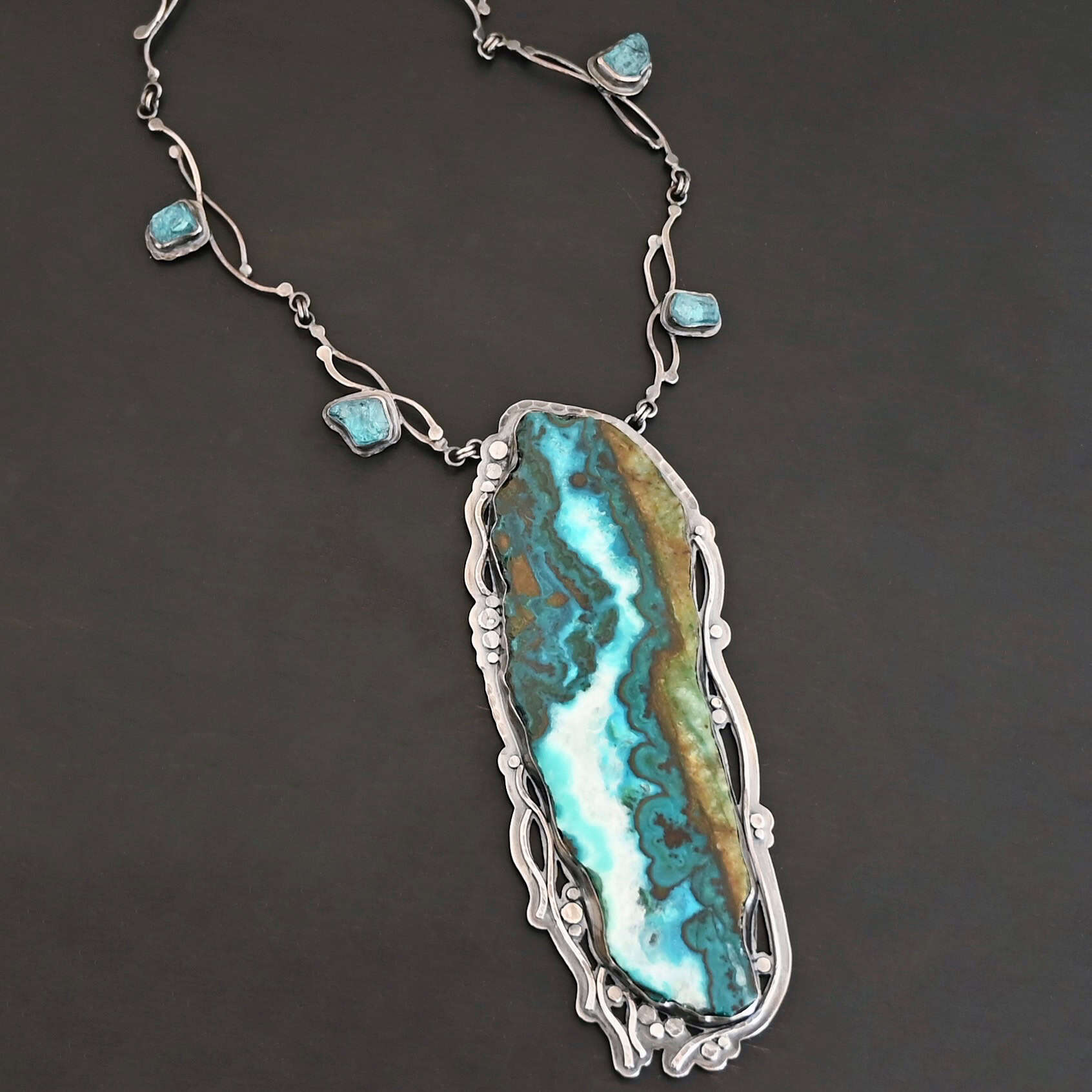 Like The River Runs Necklace Close up of the "Like The River Runs" Necklace. Made out of sterling silver, azurite, and malachite and apatite. The azurite and malachite are mixed within the middle stone. It is a mix of a dark teal, light blue, white, dark green, and light green colors. The light blue and white flow throughout the middle of the stone, and the dark and light green outline the edges of the stone, so it looks as if there is water flowing through the necklace. It is set in sterling silver. Around the setting are silver dots that are hammered so that they are flat. There are also thin silver lines on the bottom of the necklace that look as if they are flowing downwards. There are two light blue stones on the collar of the necklace, one on the left side and one on the right side. Necklace is set against a dark background.