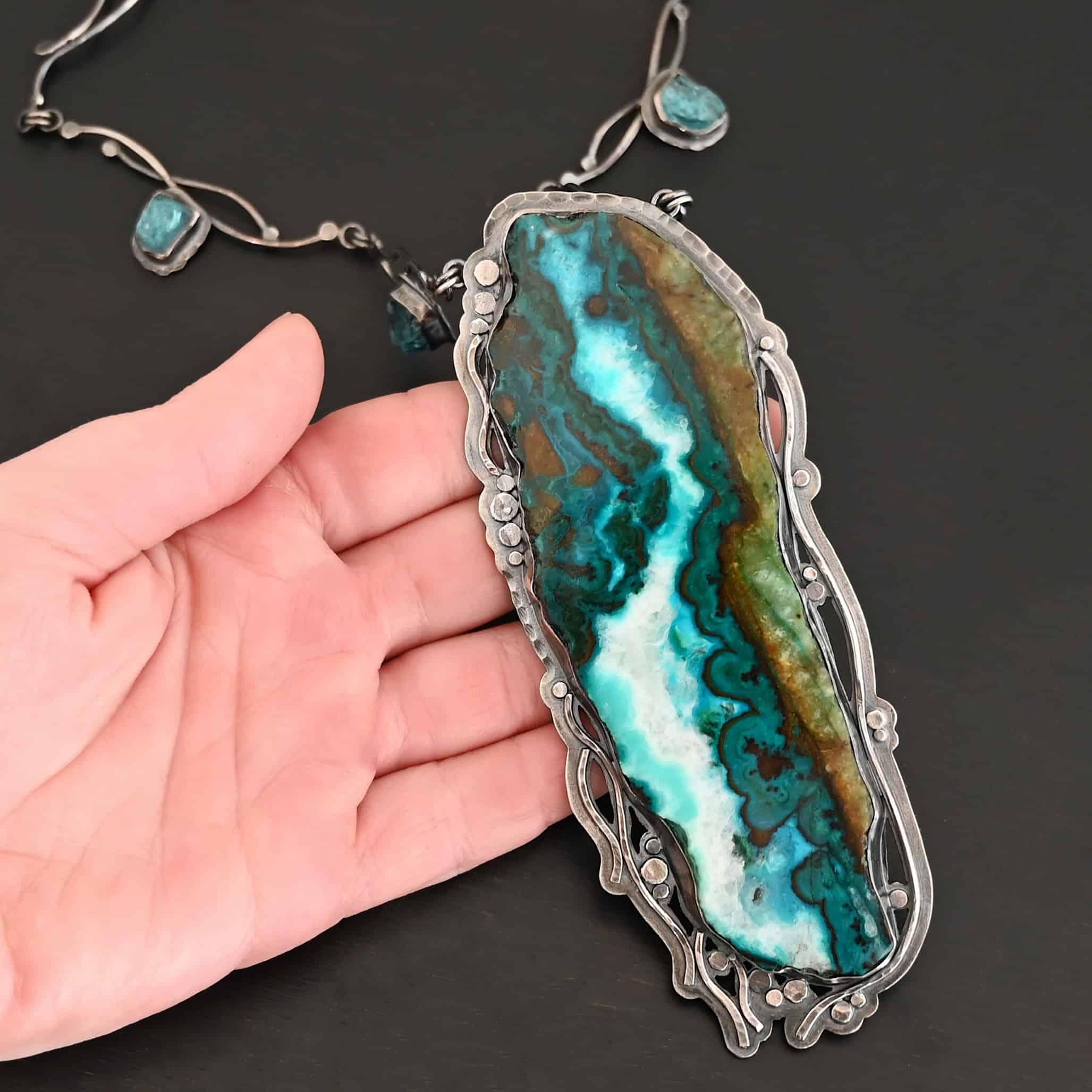 Like The River Runs Necklace New Close up of the "Like The River Runs" Necklace. Made out of sterling silver, azurite, and malachite and apatite. The azurite and malachite are mixed within the middle stone. It is a mix of a dark teal, light blue, white, dark green, and light green colors. The light blue and white flow throughout the middle of the stone, and the dark and light green outline the edges of the stone, so it looks as if there is water flowing through the necklace. It is set in sterling silver. Around the setting are silver dots that are hammered so that they are flat. There are also thin silver lines on the bottom of the necklace that look as if they are flowing downwards. There are two light blue stones on the collar of the necklace, one on the left side and one on the right side. Necklace is sitting on the fingers of a hand to demonstrate size of the pendant.