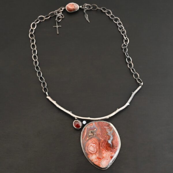 "Inner Planet" Necklace. Made out of Sterling Silver, Teepee Canyon Agate, Garnet & Aquamarine. Agate is the largest stone in a slight oval shape with a slight pointed end. It is a mix of red swirls. The aquamarine is closest to the agate. It is in the top left corner of the necklace. It is small and circular. The garnet is next to the aquamarine. It is oval shaped and a dark red. The stones are soldered to a casted sterling silver branch that is curved to go across the neck.