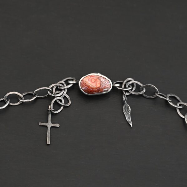 Clasp for the "Inner Planet" Necklace. There is a small, oval, red, white, and orange agate with two hooks on the back for the links to connect to, as well as a small cross and angel wing charm.