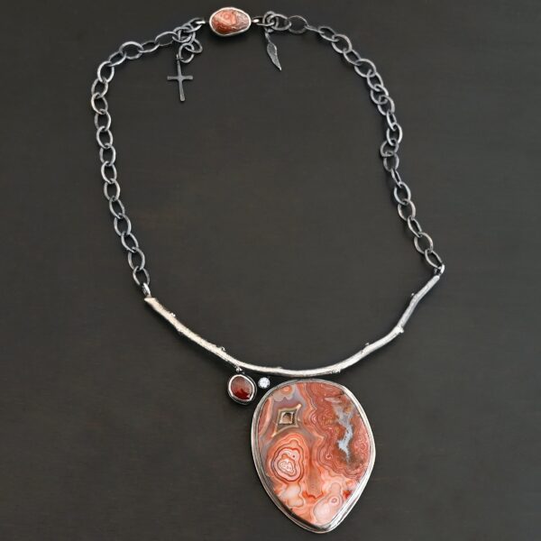 "Inner Planet" Necklace. Made out of Sterling Silver, Teepee Canyon Agate, Garnet & Aquamarine. Agate is the largest stone in a slight oval shape with a slight pointed end. It is a mix of red swirls. The aquamarine is closest to the agate. It is in the top left corner of the necklace. It is small and circular. The garnet is next to the aquamarine. It is oval shaped and a dark red. The stones are soldered to a casted sterling silver branch that is curved to go across the neck.