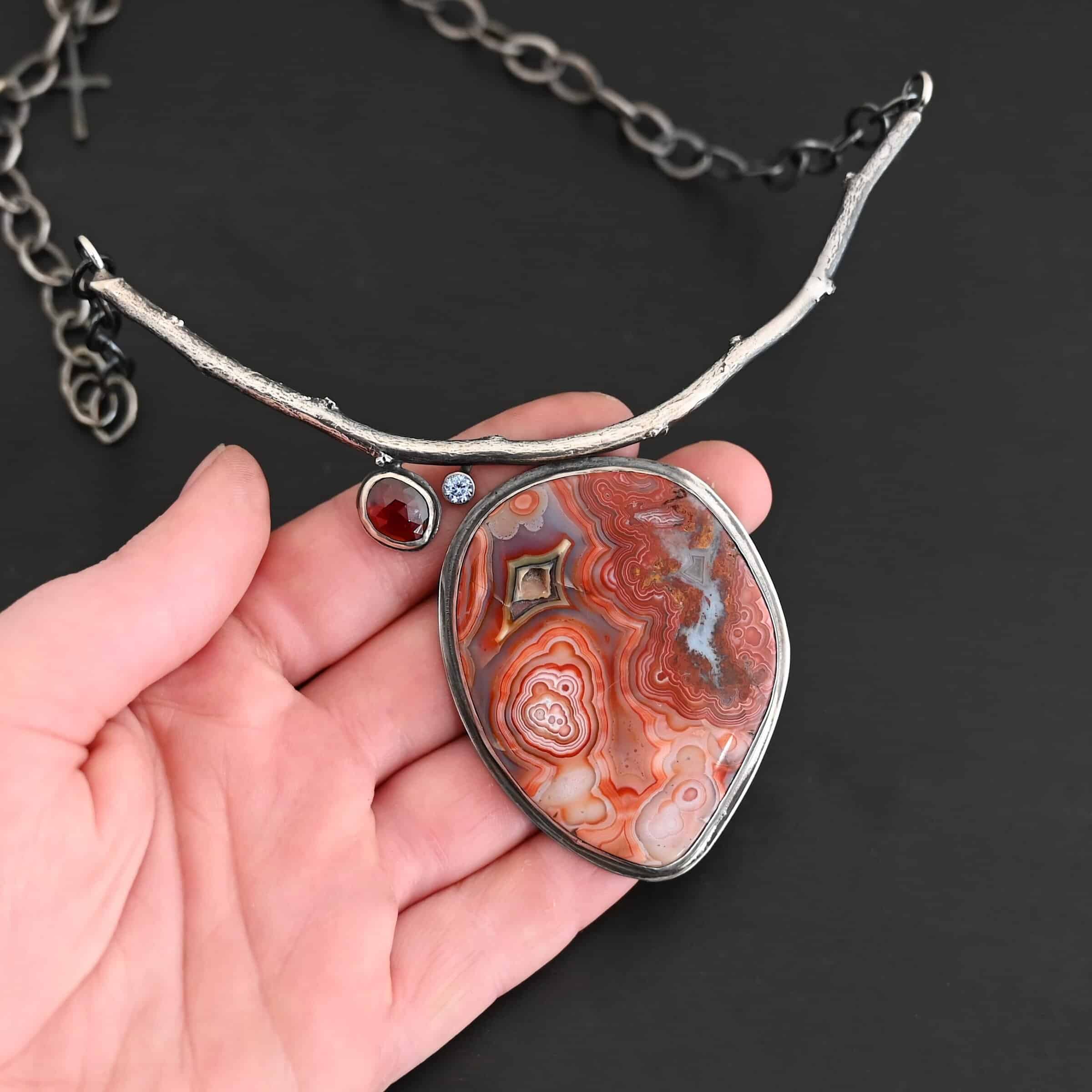 Inner Planet Necklace "Inner Planet" Necklace. Made out of Sterling Silver, Teepee Canyon Agate, Garnet & Aquamarine. Agate is the largest stone in a slight oval shape with a slight pointed end. It is a mix of red swirls. The aquamarine is closest to the agate. It is in the top left corner of the necklace. It is small and circular. The garnet is next to the aquamarine. It is oval shaped and a dark red. The stones are soldered to a casted sterling silver branch that is curved to go across the neck.