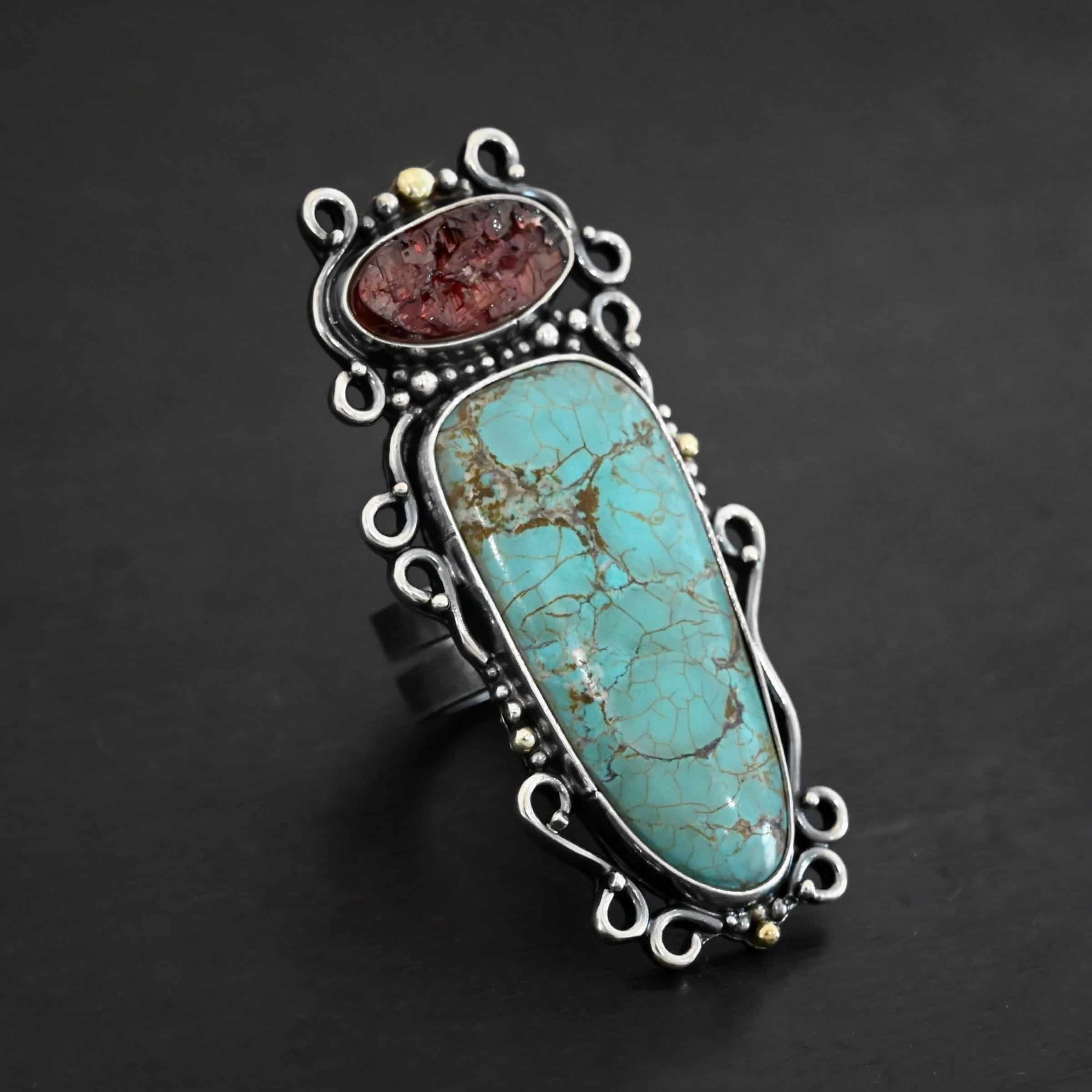 Dream Protector Ring Close up of the "Dream Protector" Ring. Made out of sterling silver, gold, turquoise, and garnet. The top stone of the ring is an oval garnet that is rough and textured. It is a dark red. The largest stone in the ring is a large oval shaped turquoise. It is set the long way up and down. It is a dark blue green color with dark green lines going throughout the entire ring. There are three gold dots on the right. One on the top and one on the bottom. There is one on the right side of the ring as well. There are silver dots scattered throughout the ring as well. There are also silver swirls that are all around the side of the ring. Ring is double banded, but bands are connected together. Ring is set against a dark background.