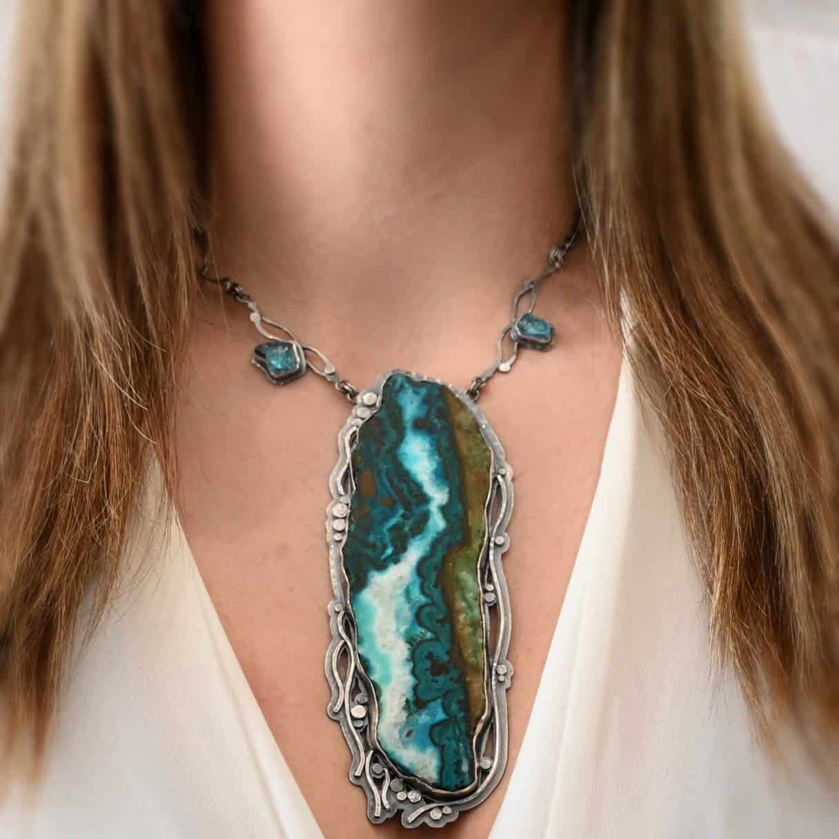 Like The River Runs Necklace Close up of the "Like The River Runs" Necklace. Made out of sterling silver, azurite, and malachite and apatite. The azurite and malachite are mixed within the middle stone. It is a mix of a dark teal, light blue, white, dark green, and light green colors. The light blue and white flow throughout the middle of the stone, and the dark and light green outline the edges of the stone, so it looks as if there is water flowing through the necklace. It is set in sterling silver. Around the setting are silver dots that are hammered so that they are flat. There are also thin silver lines on the bottom of the necklace that look as if they are flowing downwards. There are two light blue stones on the collar of the necklace, one on the left side and one on the right side. Necklace is worn by a model to demonstrate size of the pendant. Model is wearing a white v-neck shirt.