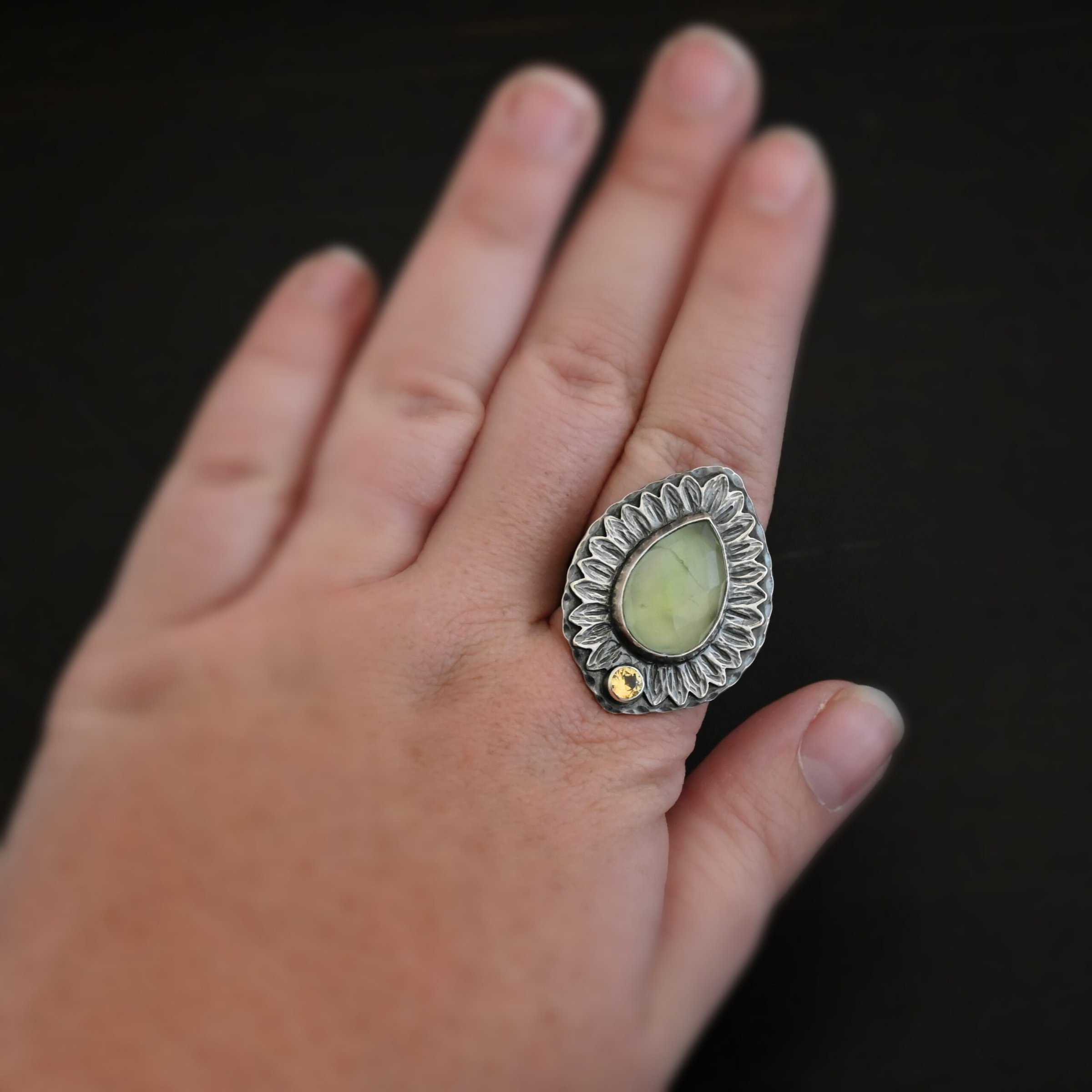 Close up of the "Summer's Flower" Ring. Made out of sterling silver, prehnite, and citrine. The prehnite is the stone in the center, and it is a teardrop shape. It is a light green color and it is rose cut. In the center of the bottom of the ring there is a small circular citrine. It is a light orange. Surrounding the stones going completely around them are sunflower looking leaves that are made out of sterling silver. This ring is oxidized. Ring is sitting on a models hand to demonstrate size of the ring.