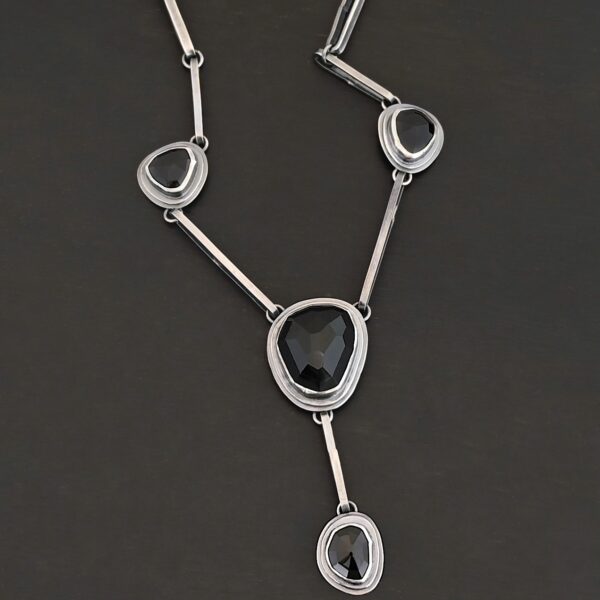 The "Mysteries Beyond" Necklace. Handmade out of sterling silver and five black onyx. Each one is set in sterling silver. There is a black onyx for the clasp of the necklace. There are four bars on each side connected by jump rings that lead to the next two black onyx, one on the left side and one on the right. Then one bar on the right and left lead down to another large black onyx. There is one more bar that leads down to the bottom of the necklace, with one more black onyx. Necklace is set against a black background.