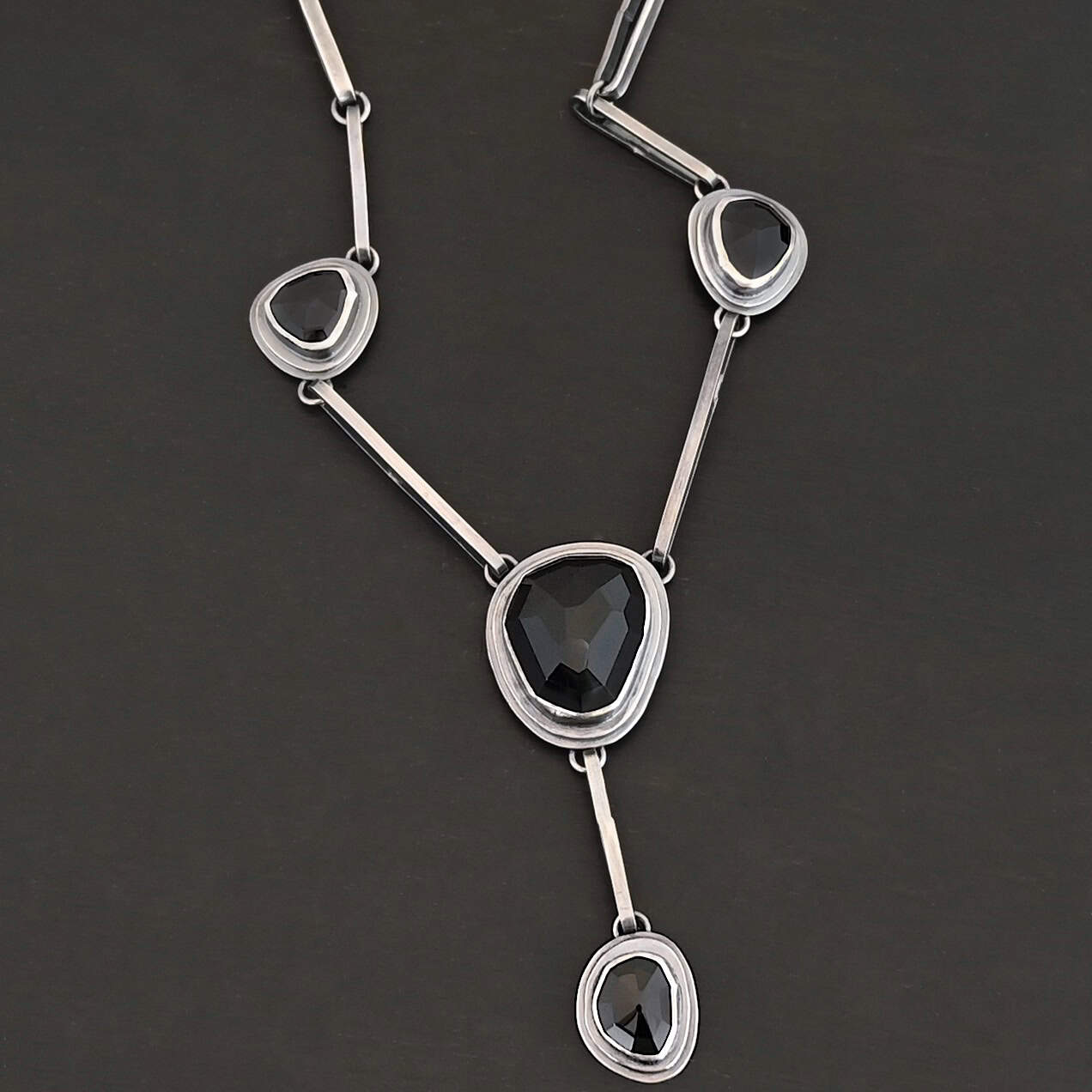 Mysteries Beyond Necklace The "Mysteries Beyond" Necklace. Handmade out of sterling silver and five black onyx. Each one is set in sterling silver. There is a black onyx for the clasp of the necklace. There are four bars on each side connected by jump rings that lead to the next two black onyx, one on the left side and one on the right. Then one bar on the right and left lead down to another large black onyx. There is one more bar that leads down to the bottom of the necklace, with one more black onyx. Necklace is set against a black background.