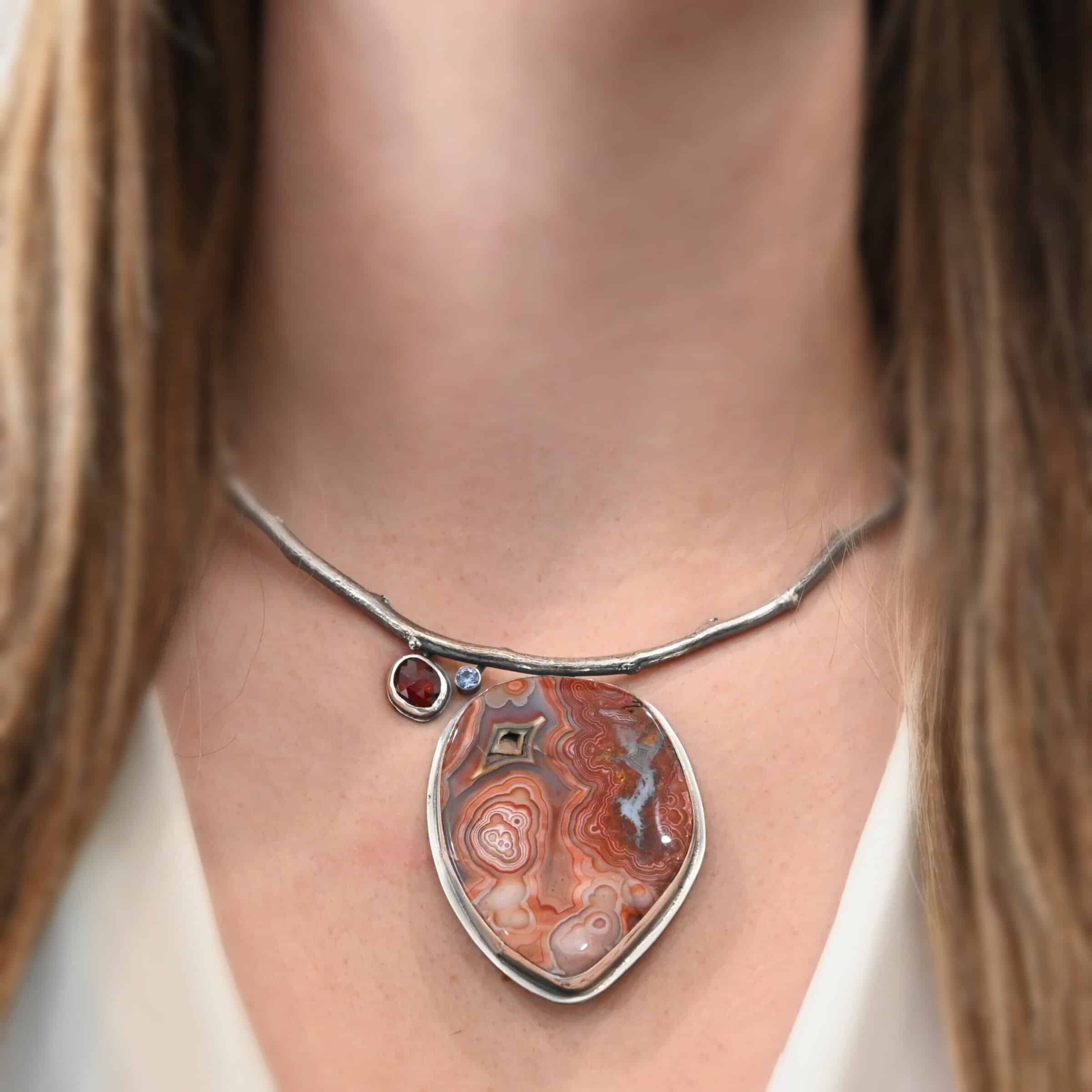Inner Planet Necklace "Inner Planet" Necklace. Made out of Sterling Silver, Teepee Canyon Agate, Garnet & Aquamarine. Agate is the largest stone in a slight oval shape with a slight pointed end. It is a mix of red swirls. The aquamarine is closest to the agate. It is in the top left corner of the necklace. It is small and circular. The garnet is next to the aquamarine. It is oval shaped and a dark red. The stones are soldered to a casted sterling silver branch that is curved to go across the neck. Model is wearing a white v-neck shirt.
