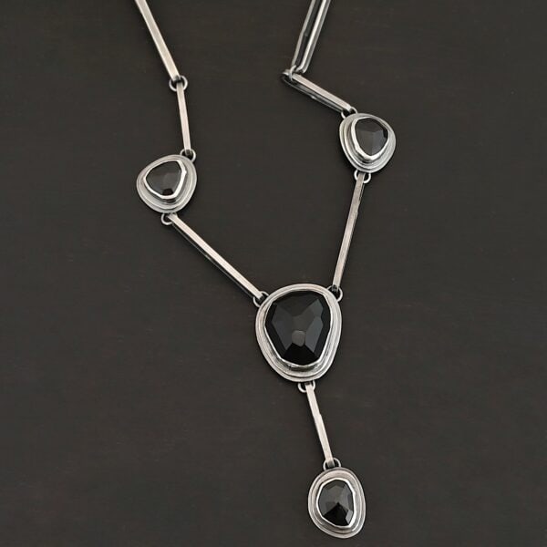 The "Mysteries Beyond" Necklace. Handmade out of sterling silver and five black onyx. Each one is set in sterling silver. There is a black onyx for the clasp of the necklace. There are four bars on each side connected by jump rings that lead to the next two black onyx, one on the left side and one on the right. Then one bar on the right and left lead down to another large black onyx. There is one more bar that leads down to the bottom of the necklace, with one more black onyx. Necklace is set against a black background.
