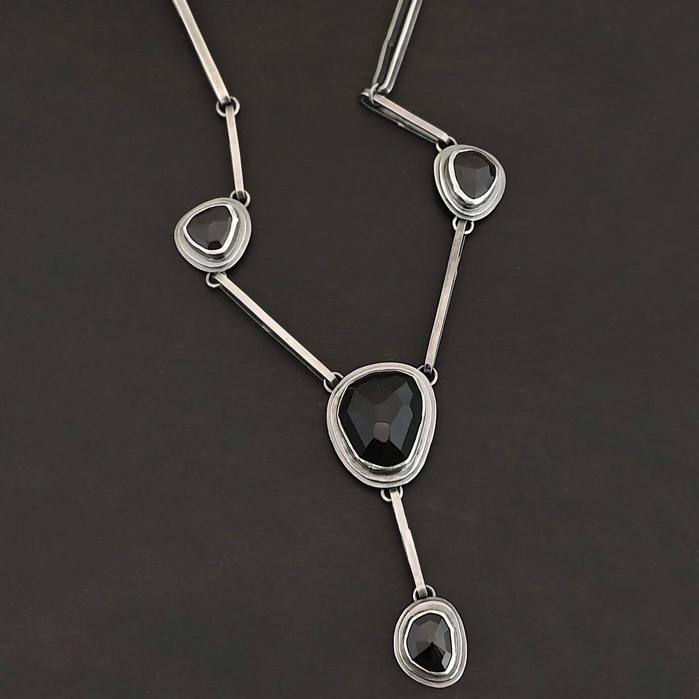 Mysteries Beyond Necklace The "Mysteries Beyond" Necklace. Handmade out of sterling silver and five black onyx. Each one is set in sterling silver. There is a black onyx for the clasp of the necklace. There are four bars on each side connected by jump rings that lead to the next two black onyx, one on the left side and one on the right. Then one bar on the right and left lead down to another large black onyx. There is one more bar that leads down to the bottom of the necklace, with one more black onyx. Necklace is set against a black background.