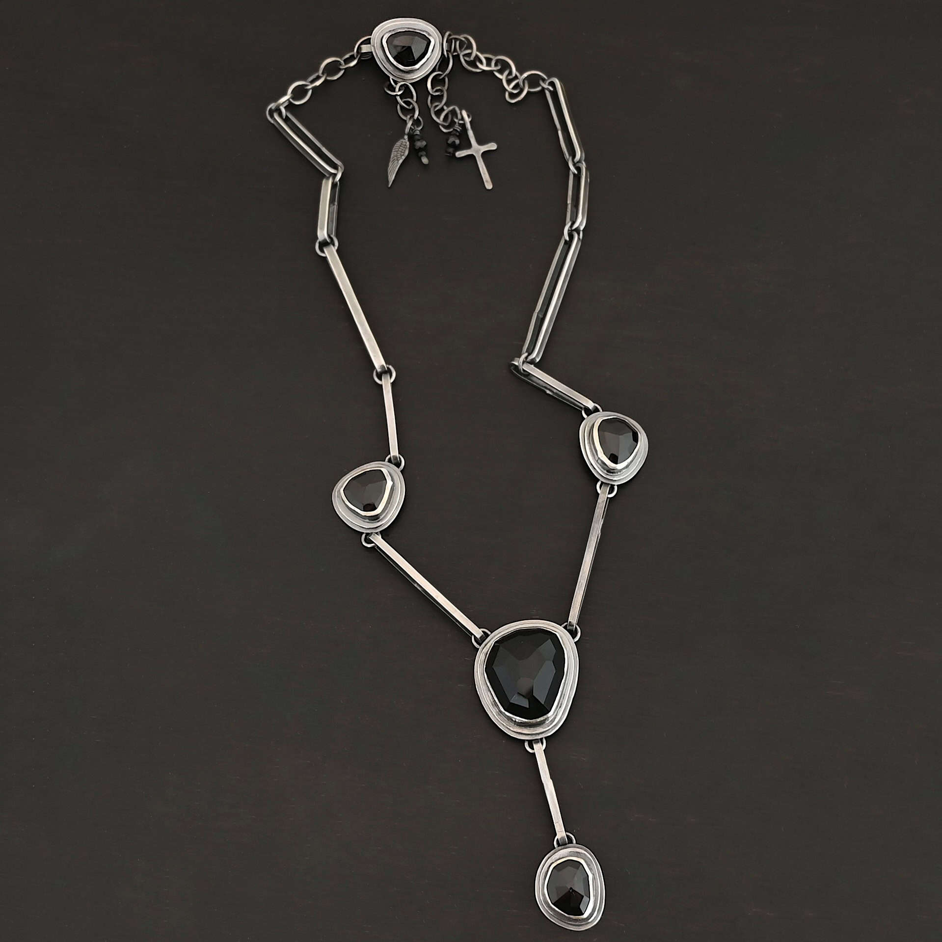 Mysteries Beyond Necklace The "Mysteries Beyond" Necklace. Handmade out of sterling silver and five black onyx. Each one is set in sterling silver. There is a black onyx for the clasp of the necklace. There are four bars on each side connected by jump rings that lead to the next two black onyx, one on the left side and one on the right. Then one bar on the right and left lead down to another large black onyx. There is one more bar that leads down to the bottom of the necklace, with one more black onyx. Necklace is set against a black background.