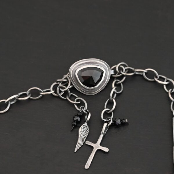 The "Mysteries Beyond" Necklace clasp. Handmade out of sterling silver and five black onyx. There is a black onyx for the clasp of the necklace. Clasp has two links on the back that each end of the chain connects to. There is a small cross and angel wing charm on the clasp. Necklace is set against a dark background.