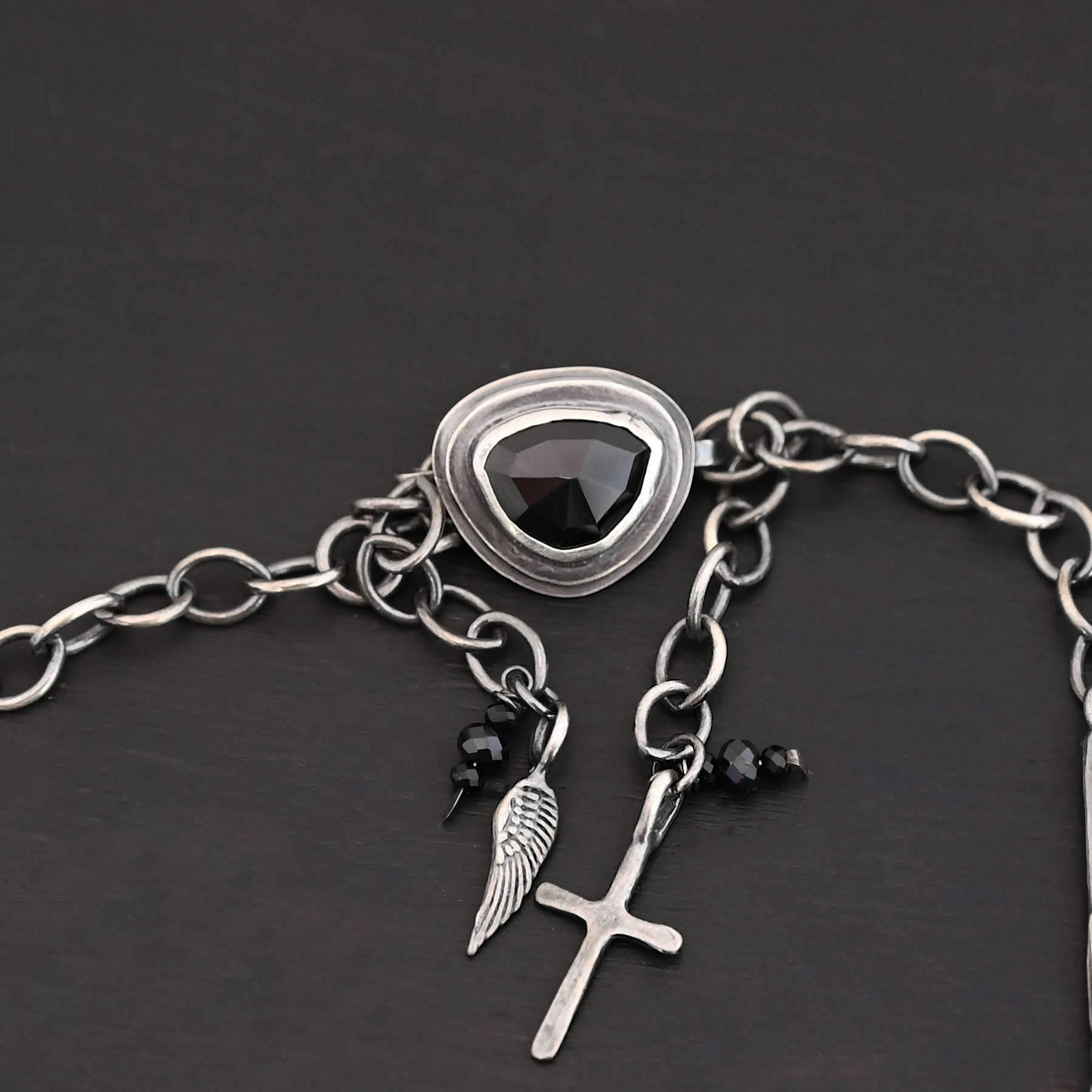 Mysteries Beyond Necklace The "Mysteries Beyond" Necklace clasp. Handmade out of sterling silver and five black onyx. There is a black onyx for the clasp of the necklace. Clasp has two links on the back that each end of the chain connects to. There is a small cross and angel wing charm on the clasp. Necklace is set against a dark background.