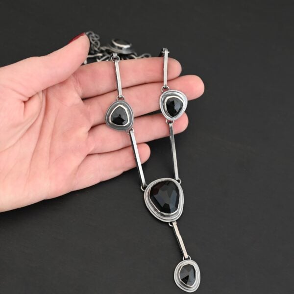 The "Mysteries Beyond" Necklace. Handmade out of sterling silver and five black onyx. Each one is set in sterling silver. There is a black onyx for the clasp of the necklace. There are four bars on each side connected by jump rings that lead to the next two black onyx, one on the left side and one on the right. Then one bar on the right and left lead down to another large black onyx. There is one more bar that leads down to the bottom of the necklace, with one more black onyx. Necklace is set against the fingertips of a hand.