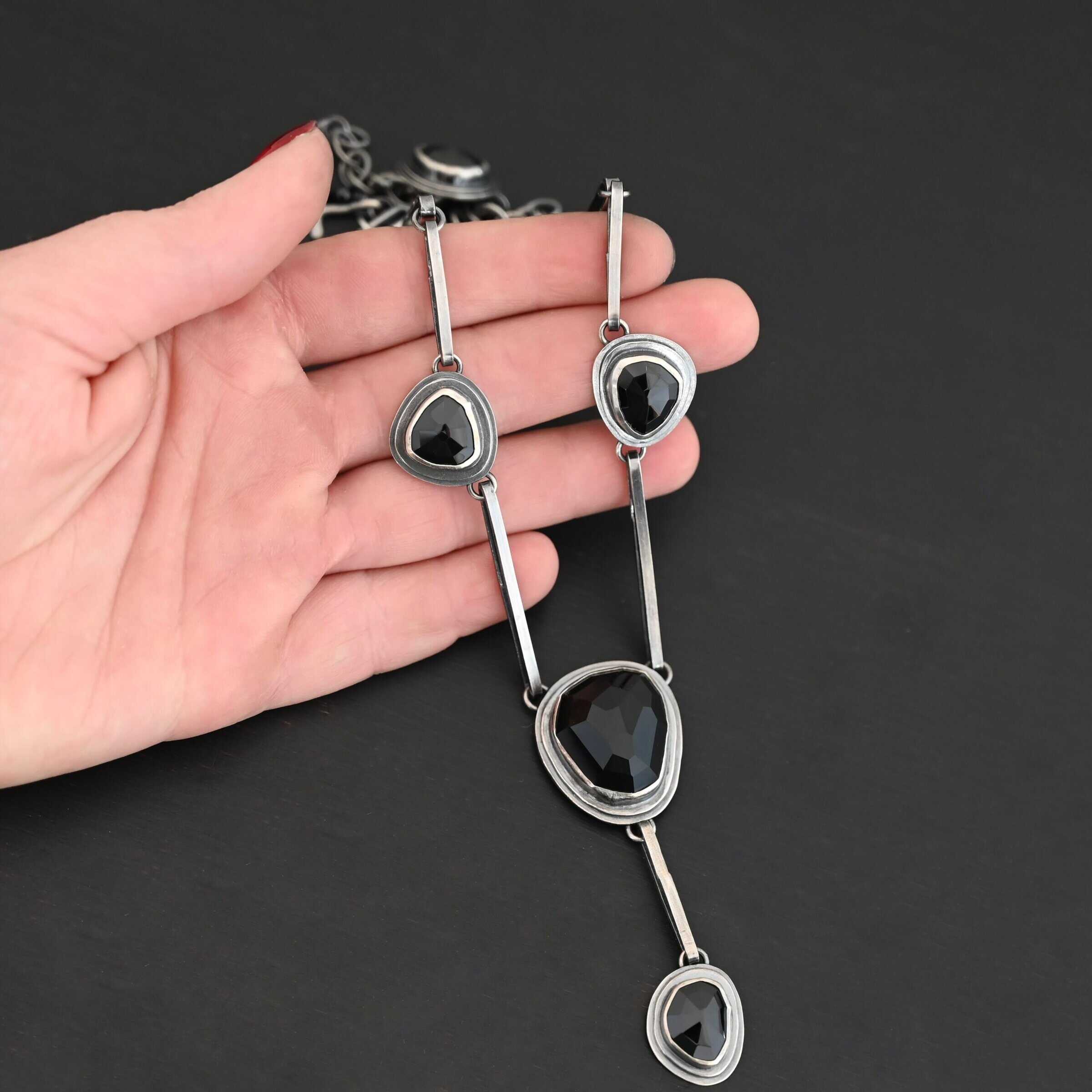 Mysteries Beyond Necklace The "Mysteries Beyond" Necklace. Handmade out of sterling silver and five black onyx. Each one is set in sterling silver. There is a black onyx for the clasp of the necklace. There are four bars on each side connected by jump rings that lead to the next two black onyx, one on the left side and one on the right. Then one bar on the right and left lead down to another large black onyx. There is one more bar that leads down to the bottom of the necklace, with one more black onyx. Necklace is set against the fingertips of a hand.