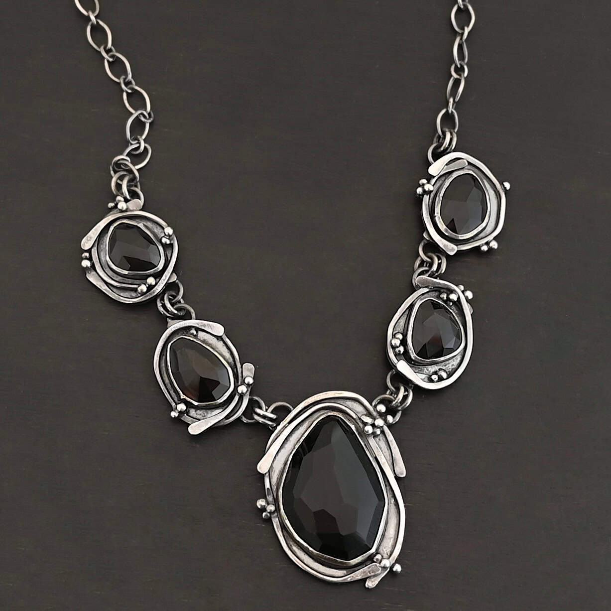 Close up of the "Blissful Beauty" Necklace. Made out of five black onyx. The center one is the biggest, with two on the left side and two more on the right. Each black onyx is set in sterling silver and has silver overlapping for each setting. Each setting also has small silver balls on it. Necklace is set against a black background.
