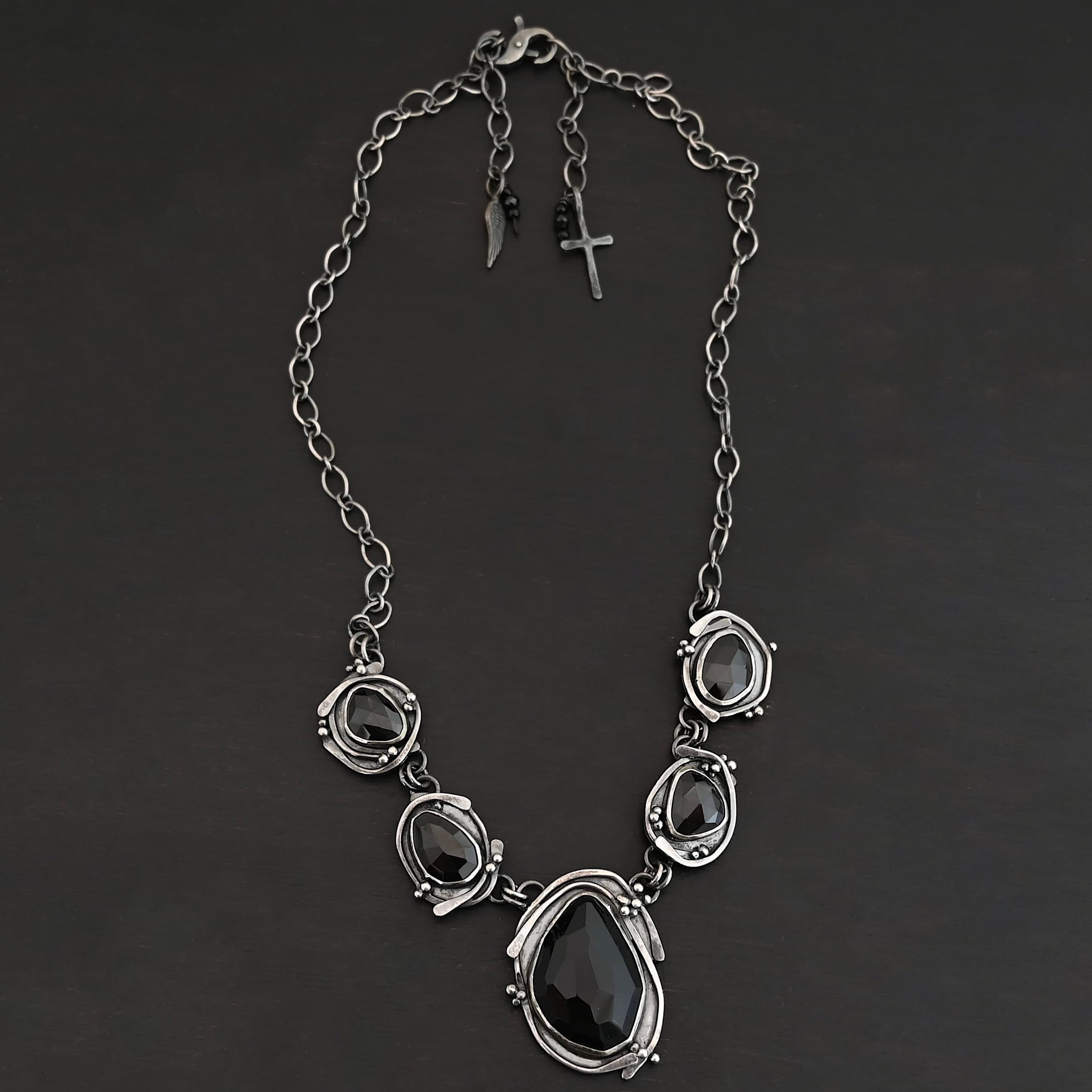 Close up of the "Blissful Beauty" Necklace. Made out of five black onyx. The center one is the biggest, with two on the left side and two more on the right. Each black onyx is set in sterling silver and has silver overlapping for each setting. Each setting also has small silver balls on it. Necklace is set against a black background.