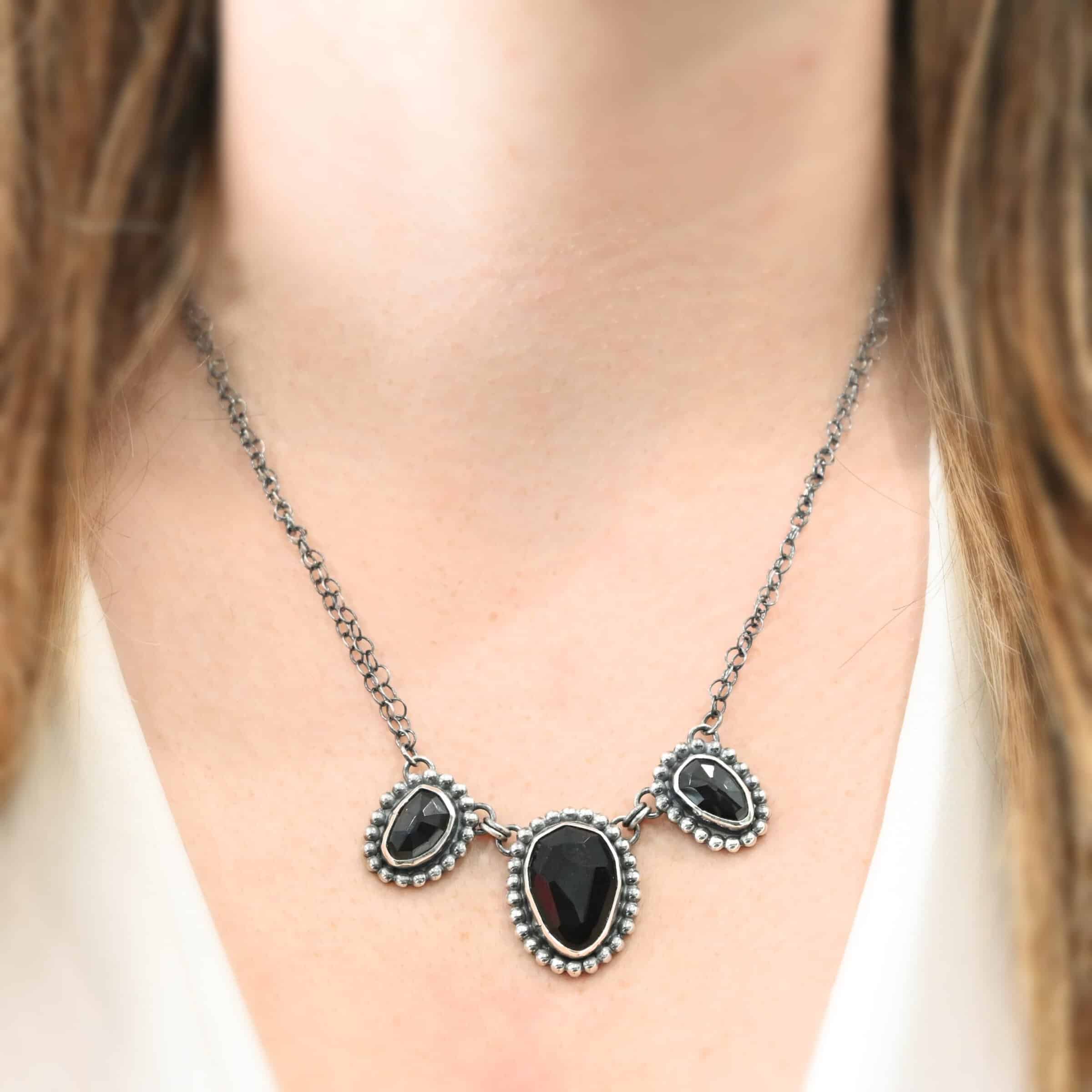 Close up of the "Escape To Tranquility" Necklace as shown on a model. Made out of sterling silver and three black onyx. The middle black onyx is the largest, with two smaller ones on the side. Each black onyx is set in silver and has silver balls going around it. Each black onyx is connected with two jump rings. Necklace is set against a dark background. Each side of the necklace is connected to two chains. Model is wearing a white v-neck shirt.