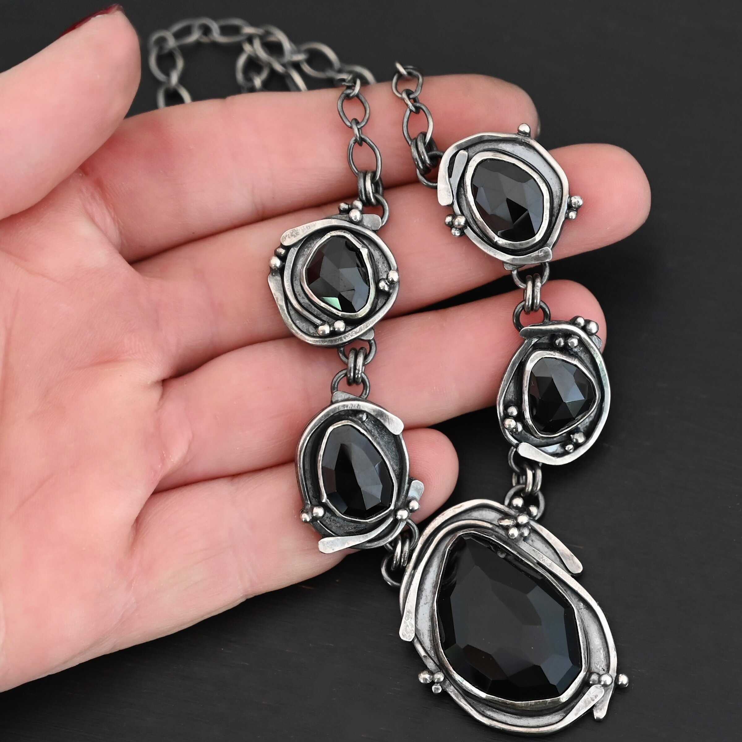 Close up of the "Blissful Beauty" Necklace. Made out of five black onyx. The center one is the biggest, with two on the left side and two more on the right. Each black onyx is set in sterling silver and has silver overlapping for each setting. Each setting also has small silver balls on it. Necklace is set against a hand.