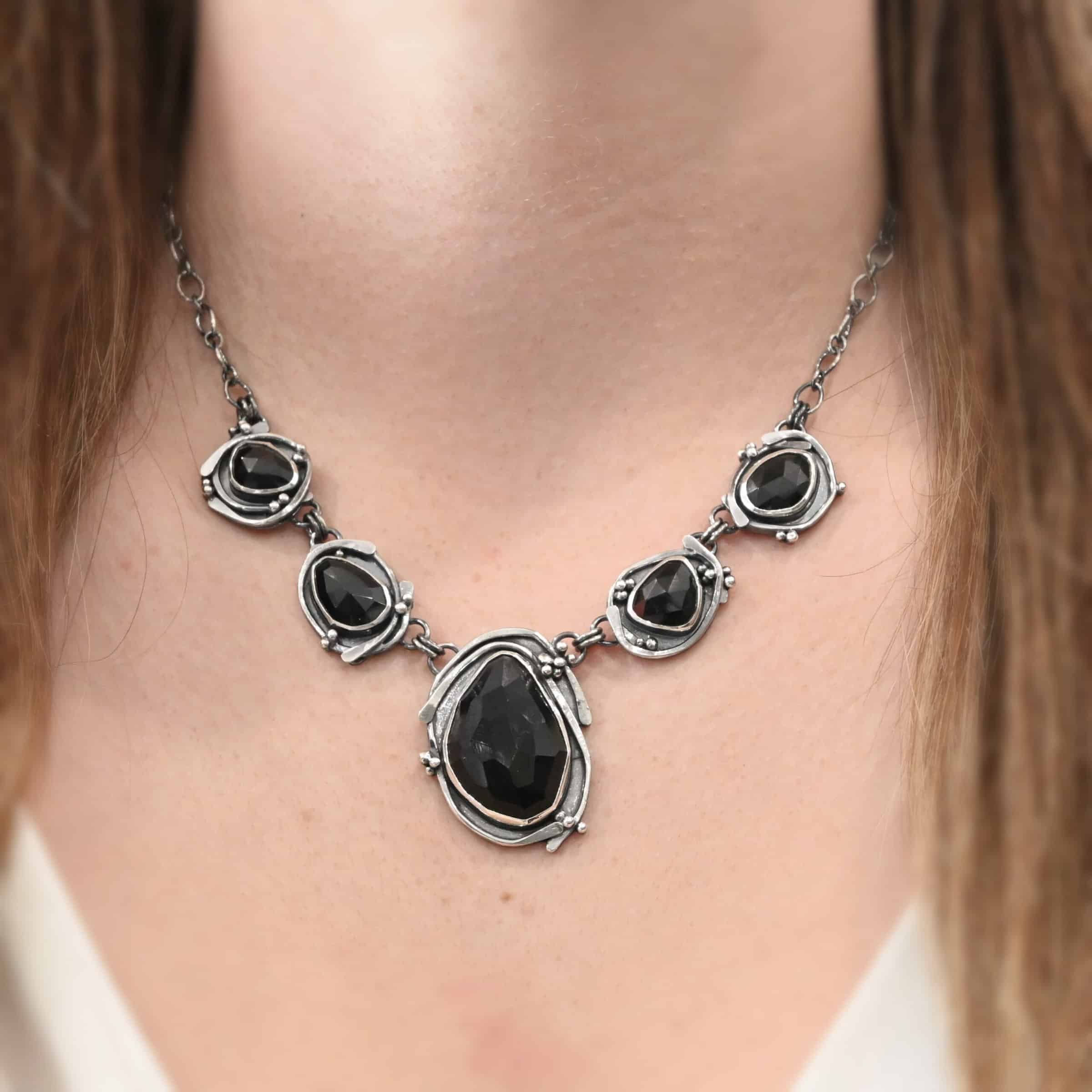 Close up of the "Blissful Beauty" Necklace as shown on a model. Made out of five black onyx. The center one is the biggest, with two on the left side and two more on the right. Each black onyx is set in sterling silver and has silver overlapping for each setting. Each setting also has small silver balls on it. Model is wearing a white v-neck shirt.
