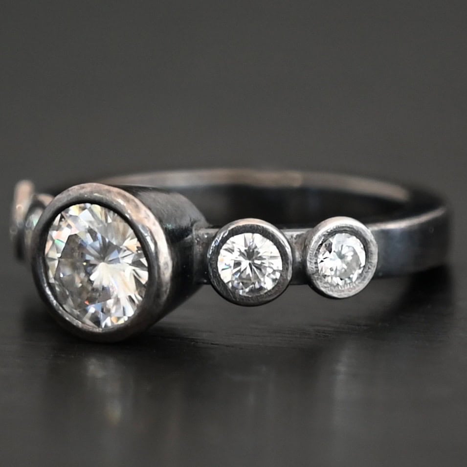 Photo of the "Heart Of Silver" Ring. There is a large moissanite in the center of the ring, with two smaller moissanites on each side of it, making for five total moissanites on the ring. Ring is set against a dark background.
