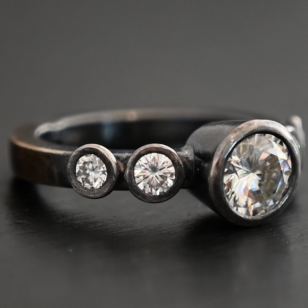 Photo of the "Heart Of Silver" Ring. There is a large moissanite in the center of the ring, with two smaller moissanites on each side of it, making for five total moissanites on the ring. Ring is set against a dark background.