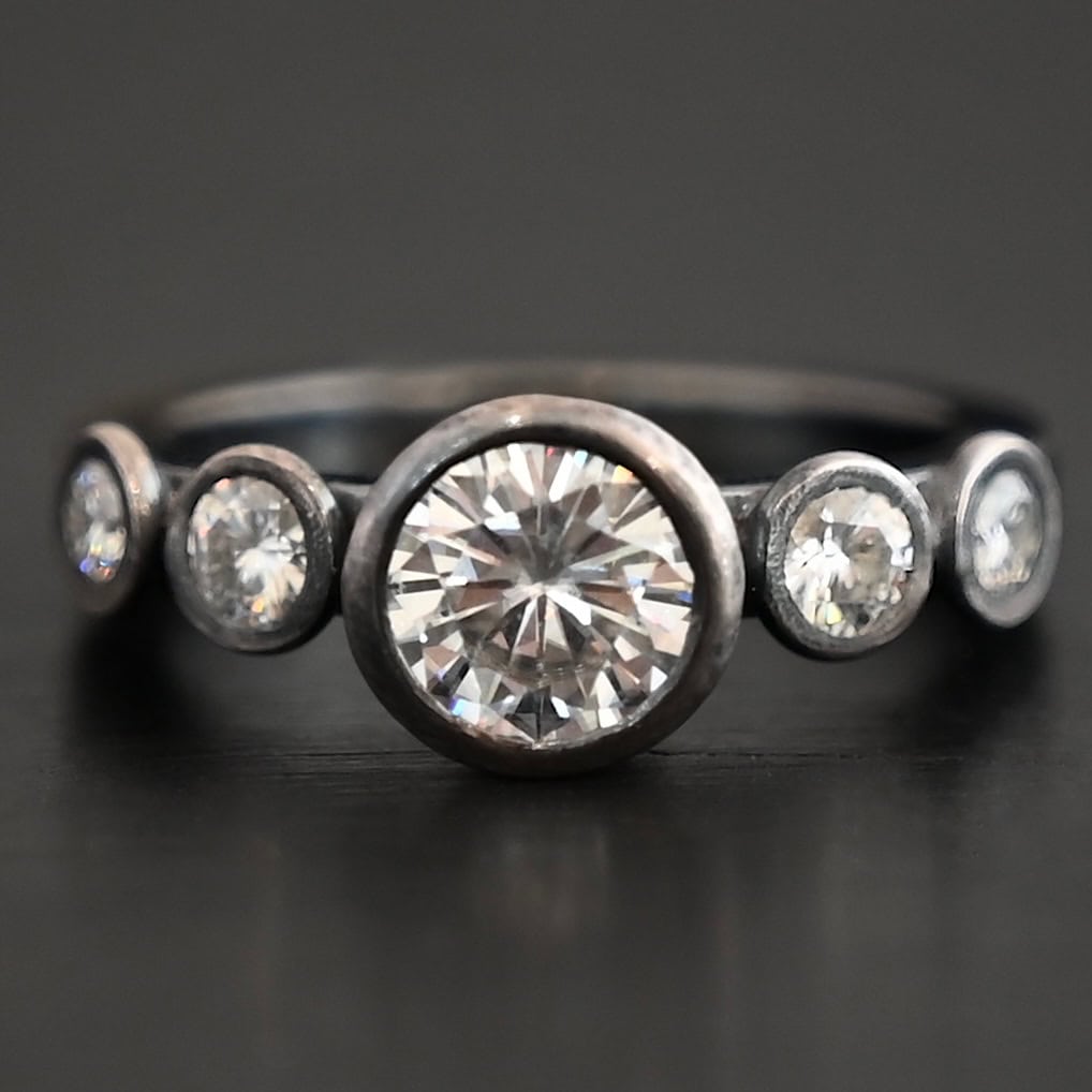 Photo of the "Heart Of Silver" Ring. There is a large moissanite in the center of the ring, with two smaller moissanites on each side of it, making for five total moissanites on the ring. Ring is set against a dark background.