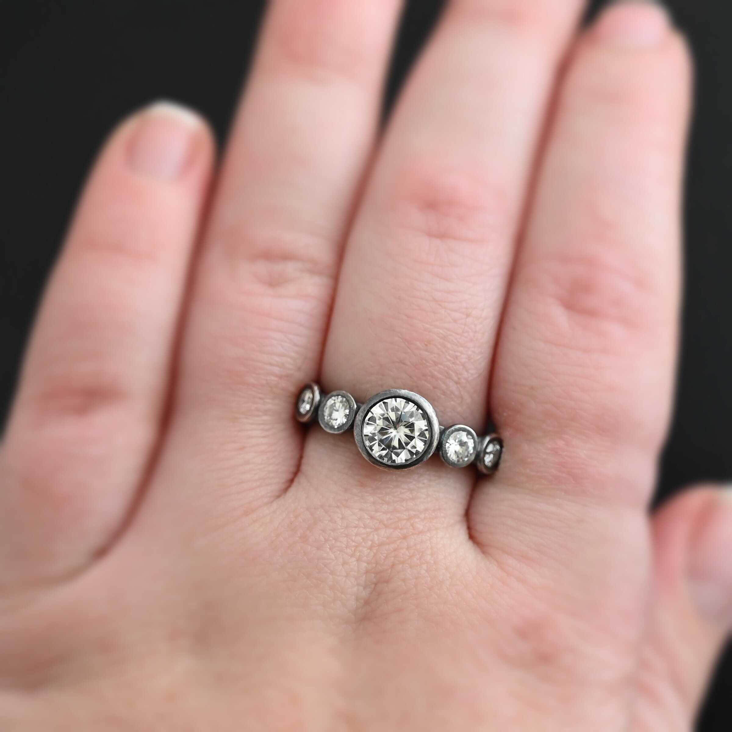 Photo of the "Heart Of Silver" Ring. There is a large moissanite in the center of the ring, with two smaller moissanites on each side of it, making for five total moissanites on the ring. Ring is worn by a model.