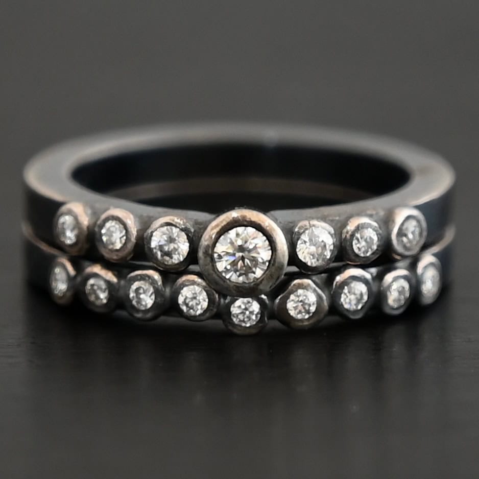 New Aura Chic Ring "Aura Chic" Ring as shown up close. Made out of 16 moissanites. There are two bands. The top band contains 7 moissanites. It also contains the largest moissanite, with three on the left side of that and three on the right. The bottom band contains 9 moissanites, all the same size. Ring is set against a dark background.