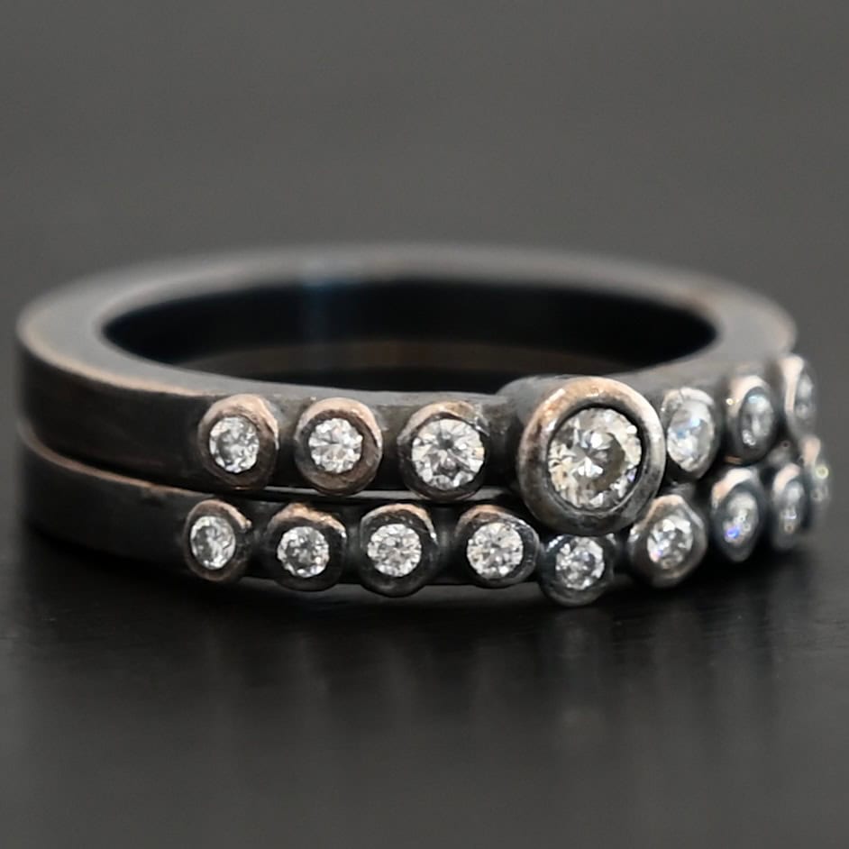 New Aura Chic Ring "Aura Chic" Ring as shown up close. Made out of 16 moissanites. There are two bands. The top band contains 7 moissanites. It also contains the largest moissanite, with three on the left side of that and three on the right. The bottom band contains 9 moissanites, all the same size. Ring is set against a dark background.