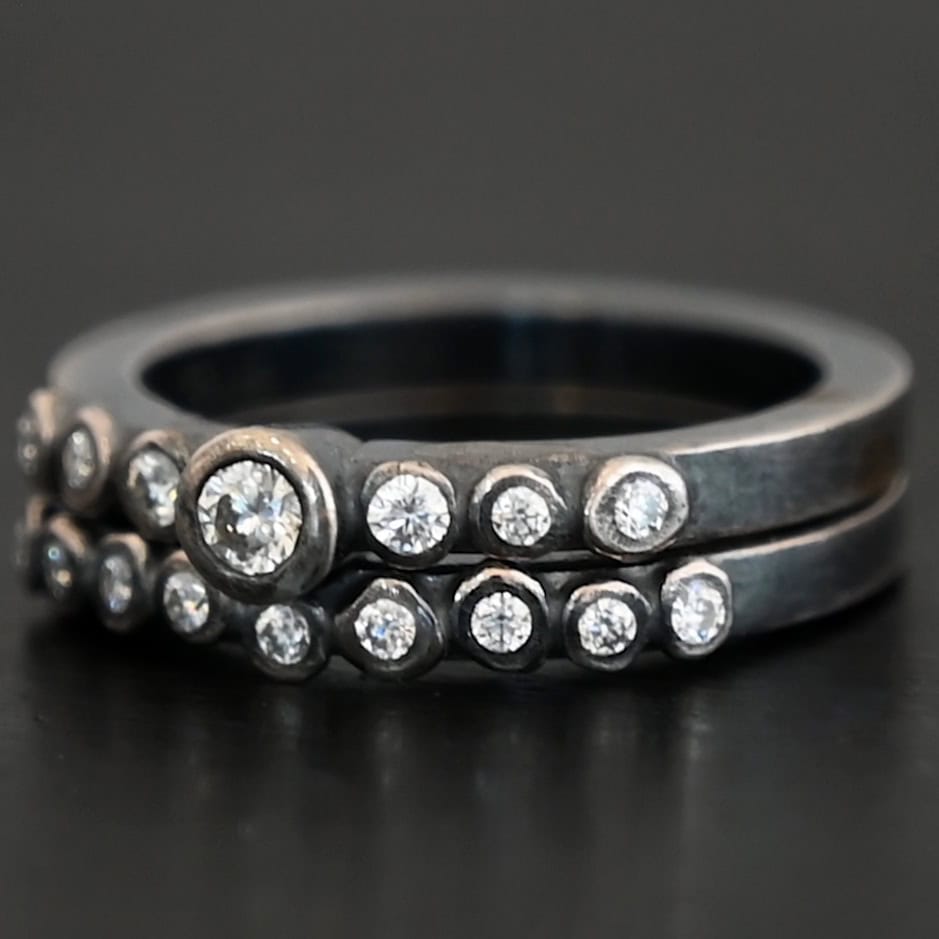 New Aura Chic Ring "Aura Chic" Ring as shown up close. Made out of 16 moissanites. There are two bands. The top band contains 7 moissanites. It also contains the largest moissanite, with three on the left side of that and three on the right. The bottom band contains 9 moissanites, all the same size. Ring is set against a dark background.