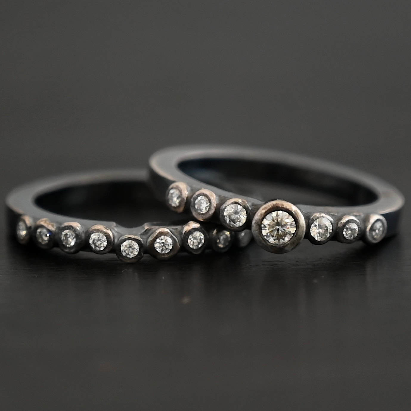 New Aura Chic Ring "Aura Chic" Ring as shown up close. Made out of 16 moissanites. There are two bands. The top band contains 7 moissanites. It also contains the largest moissanite, with three on the left side of that and three on the right. The bottom band contains 9 moissanites, all the same size. Ring is set against a dark background.