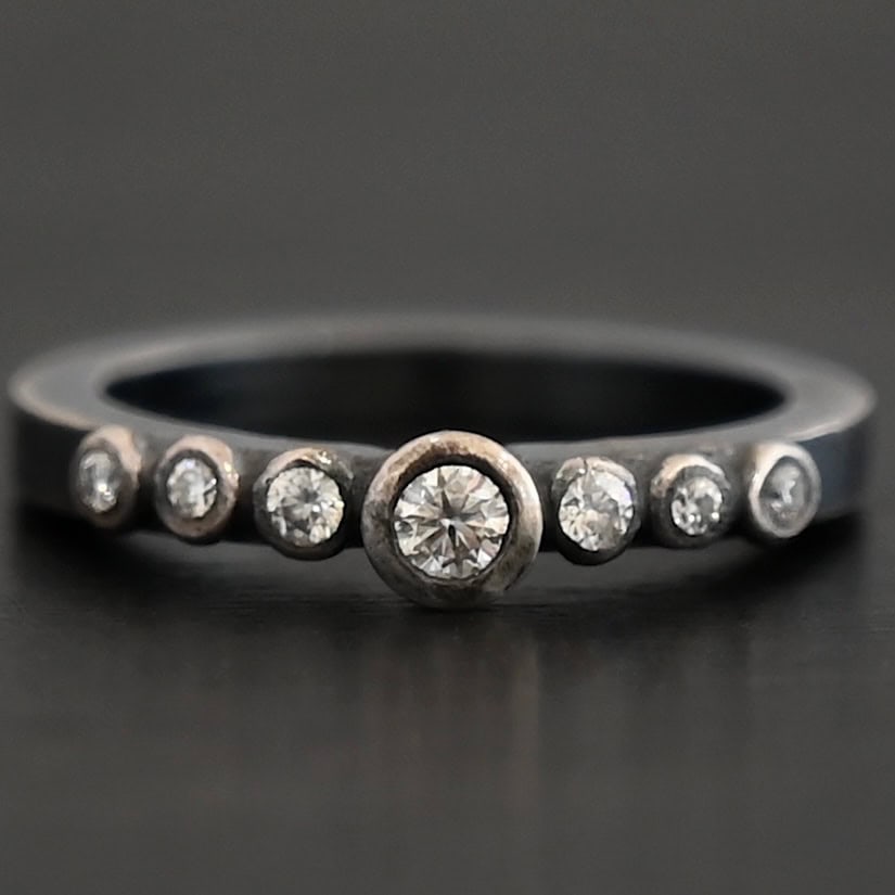 New Aura Chic Ring "Aura Chic" Ring as shown up close as one band. The top band contains 7 moissanites. It also contains the largest moissanite, with three on the left side of that and three on the right. Ring is set against a dark background.