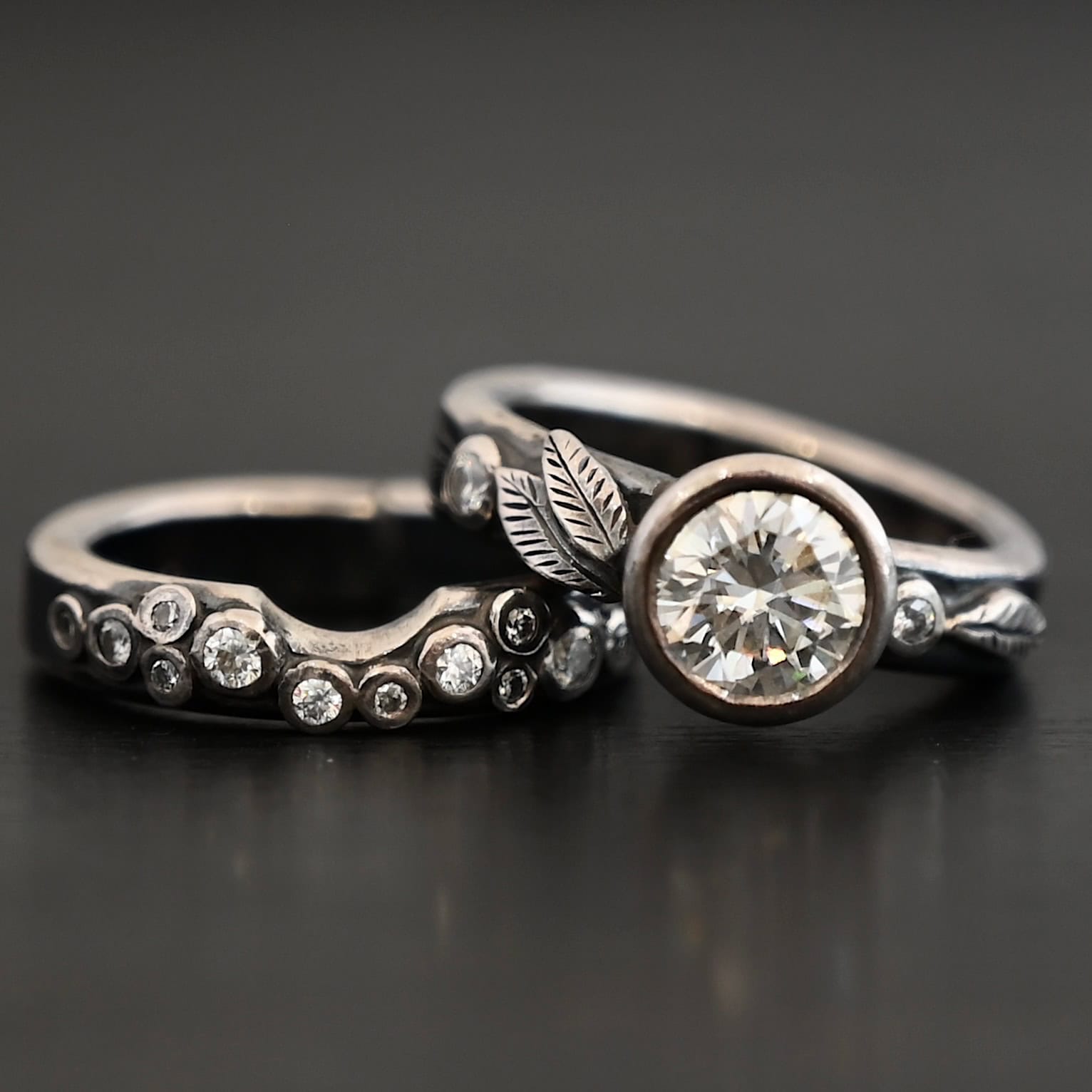 New Stars In The Sky "Stars In The Sky" Ring as shown up close. Made out of fifteen moissanites. The largest one is in the center on the top band. There is a small one on the right and another small one on the left side of the top band. There are three small engraved silver leaves on the top band, with two on the left and one on the right. The bottom band has twelve moissanites on it, all scattered. Ring is set against a dark background.