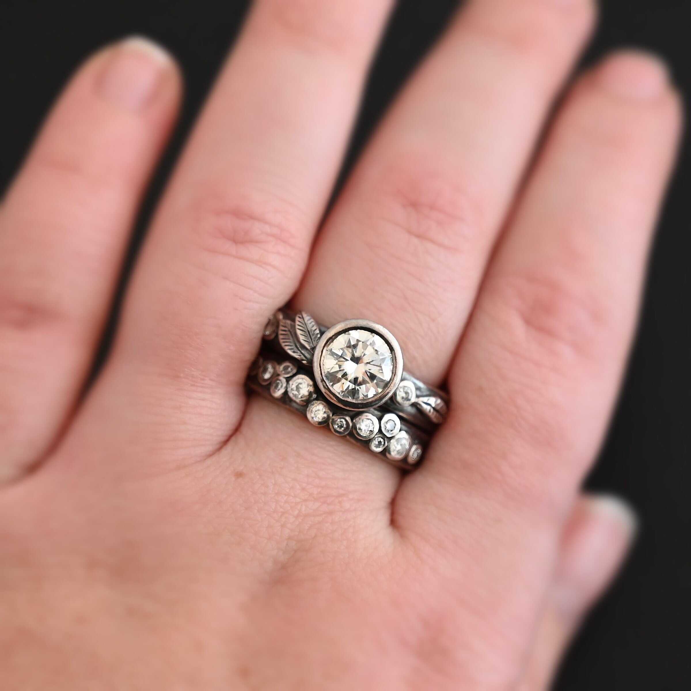 New Stars In The Sky "Stars In The Sky" Ring as shown up close. Made out of fifteen moissanites. The largest one is in the center on the top band. There is a small one on the right and another small one on the left side of the top band. There are three small engraved silver leaves on the top band, with two on the left and one on the right. The bottom band has twelve moissanites on it, all scattered. Ring is worn by a model up close.