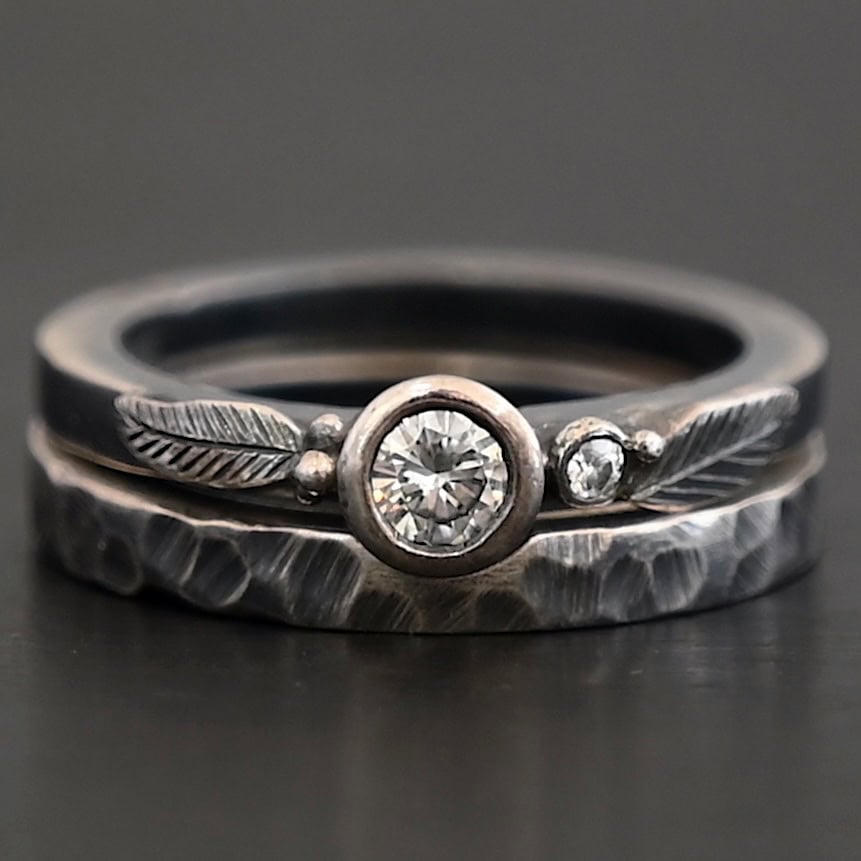 Close up of the "Shining Light" Ring. Made out of sterling silver and two moissanites. There is a larger moissanite in the center of the ring with one on the right side of that. The ring comes with two separate bands. There are two sterling silver engraved leaves on the top band, and small silver dots scattered around. The bottom band is hammered and oxidized. Ring is set against a dark background.