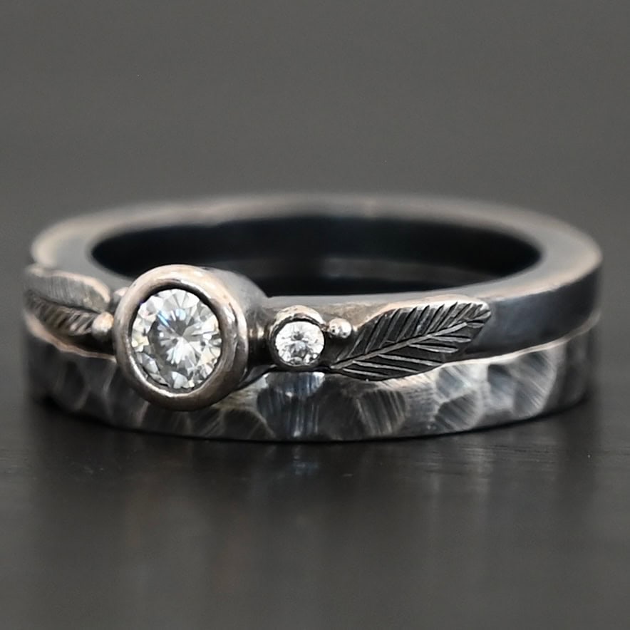 Close up of the "Shining Light" Ring. This ring is handmade out of sterling silver and moissanites. There are two moissanites ~ one as the center stone, and one on the right side of that. The ring has two separate bands that go together, as the top one sits on the bottom one in a groove. There are two silver leaves, two on the top band on each side of the center moissanite. The bottom band is hammered and oxidized silver. Ring is set against a dark background.