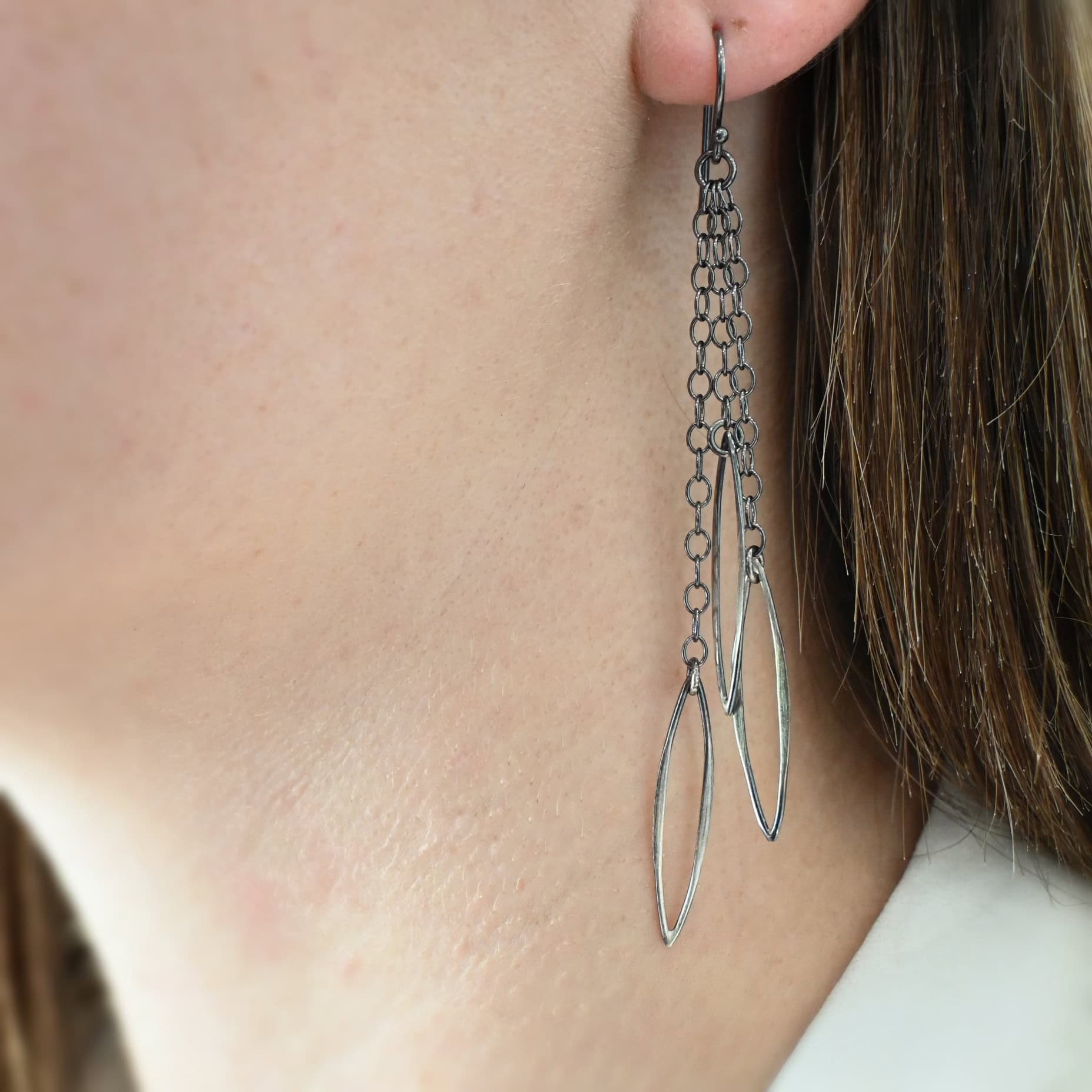 Trickling Rain Earrings Close up of the "Trickling Rain" Earrings. Made out of sterling silver. Earrings come with french hook ear wires that are sterling silver. Each earring has three strands of chain. On the end of each of these chain strands is a piece of sterling silver that is shaped like an oval, but with pointed ends on each side. Earrings are being worn on a models ear to demonstrate length and how dangly they are.