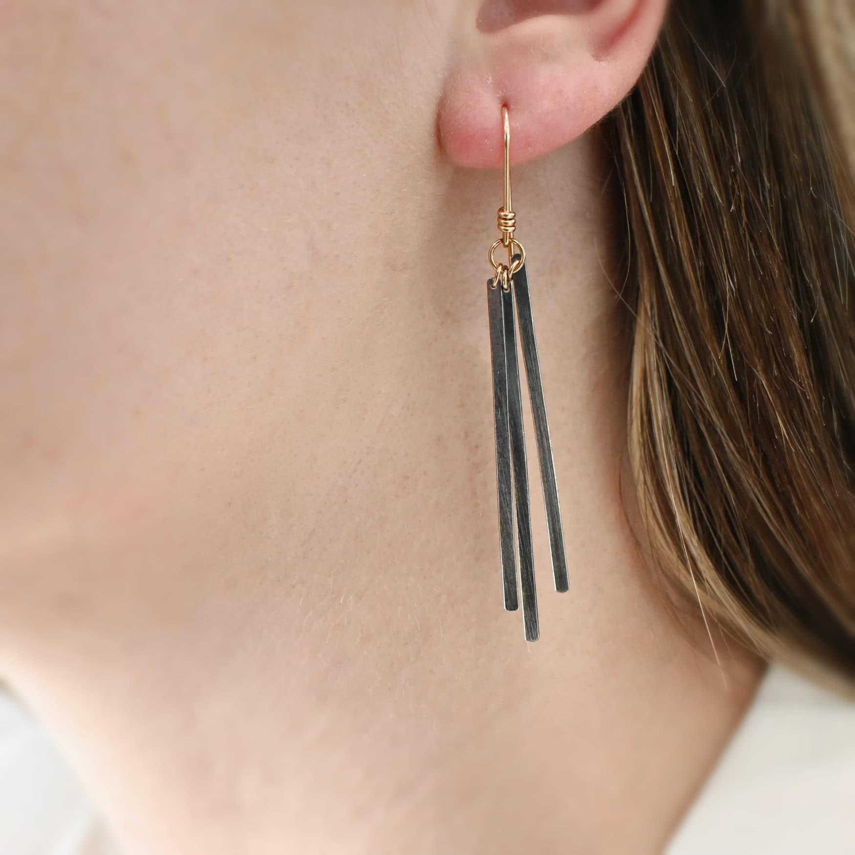 Close up of the "Moonbeam" Earrings. Made out of oxidized sterling silver and gold filled french hook ear wires. There are three slim rectangular bars of oxidized sterling silver on each earring that are connected to gold filled ear wires. Earrings are being worn by a model to demonstrate length and how dangly they are.