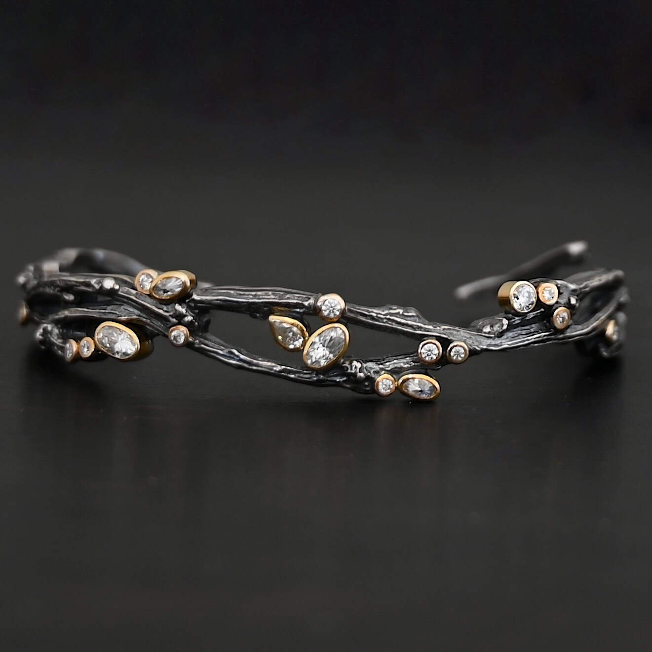 New Beautiful Dreams Cuff Close up of the "Beautiful Dreams" Cuff. Made out of sterling silver, gold, and moissanites. There are moissanites set in gold scattered throughout the bracelet. The cuff of the bracelet is made out of sterling silver thick casted twig branches. The branches flow up and down, and intertwine with one another, and the moissanites are set on and in between the branches. Cuff is set against a dark background.