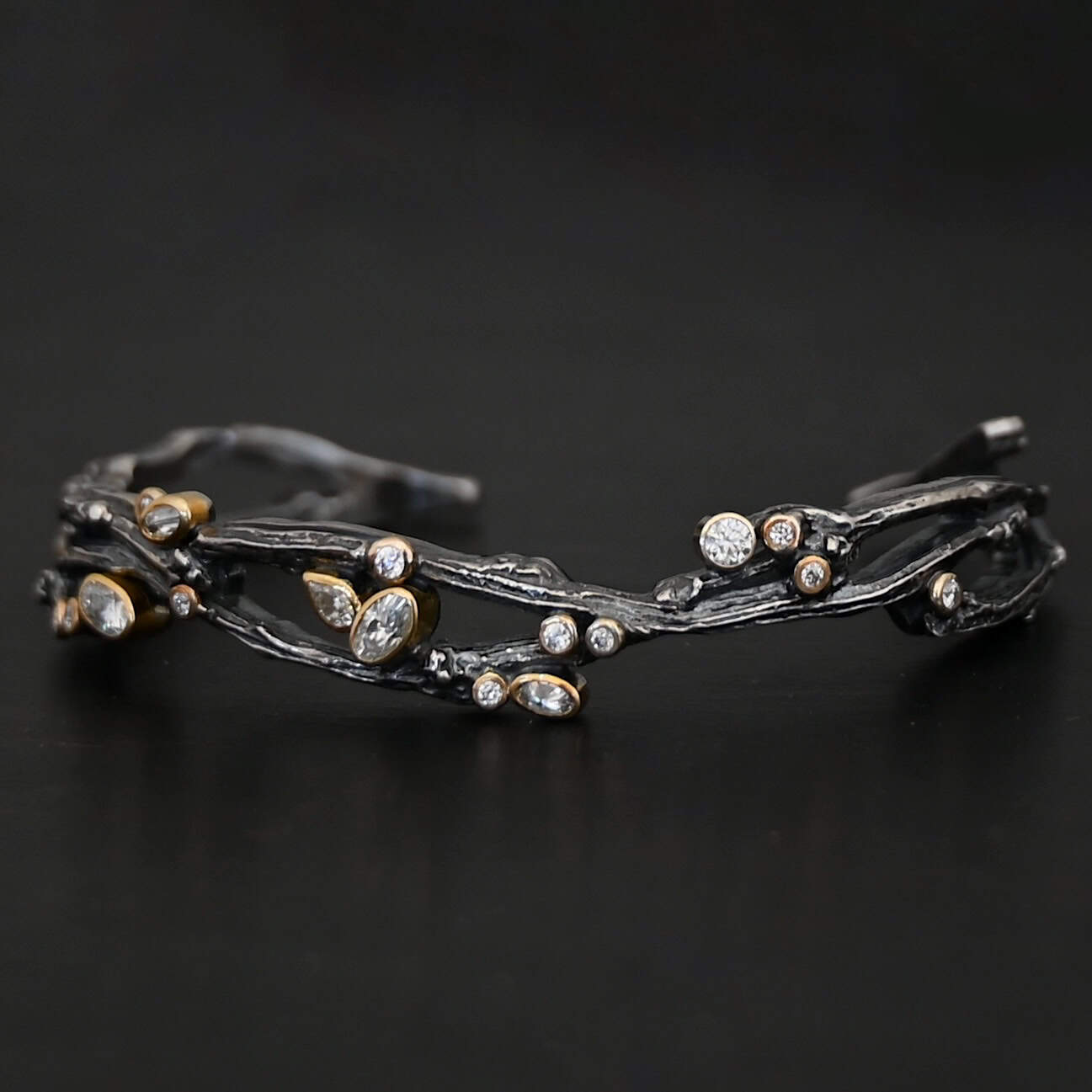 New Beautiful Dreams Cuff Close up of the "Beautiful Dreams" Cuff. Made out of sterling silver, gold, and moissanites. There are moissanites set in gold scattered throughout the bracelet. The cuff of the bracelet is made out of sterling silver thick casted twig branches. The branches flow up and down, and intertwine with one another, and the moissanites are set on and in between the branches. Cuff is set against a dark background.