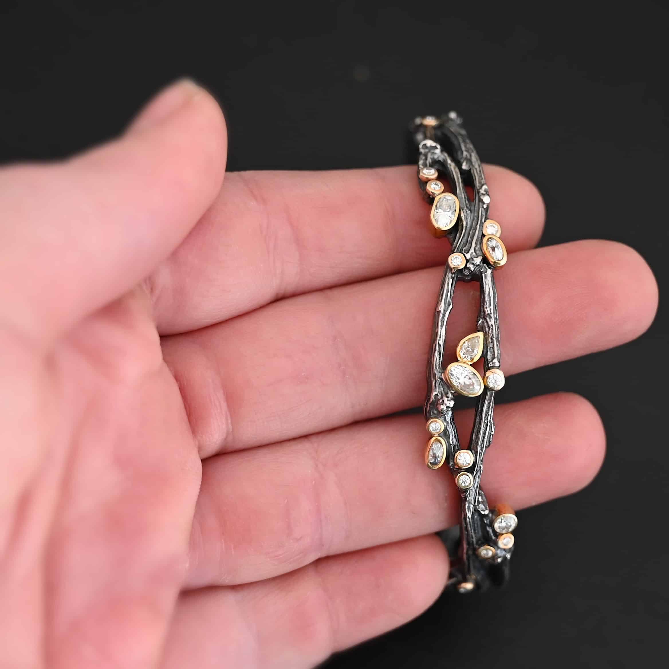 New Beautiful Dreams Cuff Close up of the "Beautiful Dreams" Cuff. Made out of sterling silver, gold, and moissanites. There are moissanites set in gold scattered throughout the bracelet. The cuff of the bracelet is made out of sterling silver thick casted twig branches. The branches flow up and down, and intertwine with one another, and the moissanites are set on and in between the branches. Cuff is set against the fingers of a hand to demonstrate thickness of the bracelet.