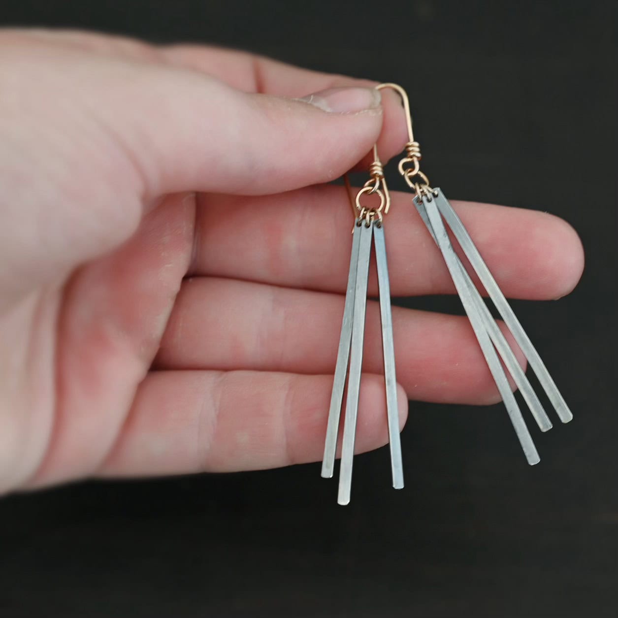 Close up of the "Moonbeam" Earrings. Made out of oxidized sterling silver and gold filled french hook ear wires. There are three slim rectangular bars of oxidized sterling silver on each earring that are connected to gold filled ear wires. Earrings are held in the fingertips to demonstrate length.