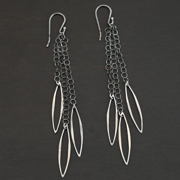 Close up of the "Trickling Rain" Earrings. Made out of sterling silver. Earrings come with french hook ear wires that are sterling silver. Each earring has three strands of chain. On the end of each of these chain strands is a piece of sterling silver that is shaped like an oval, but with pointed ends on each side.