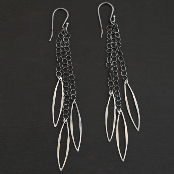 Close up of the "Trickling Rain" Earrings. Made out of sterling silver. Earrings come with french hook ear wires that are sterling silver. Each earring has three strands of chain. On the end of each of these chain strands is a piece of sterling silver that is shaped like an oval, but with pointed ends on each side.