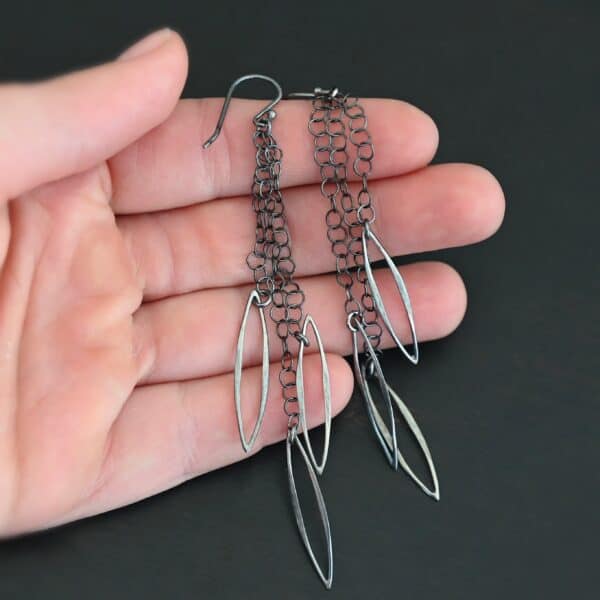 Close up of the "Trickling Rain" Earrings. Made out of sterling silver. Earrings come with french hook ear wires that are sterling silver. Each earring has three strands of chain. On the end of each of these chain strands is a piece of sterling silver that is shaped like an oval, but with pointed ends on each side.