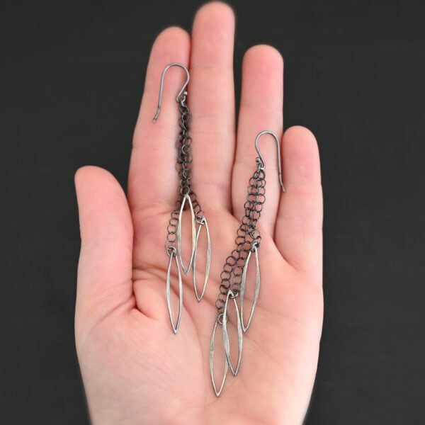 Close up of the "Trickling Rain" Earrings. Made out of sterling silver. Earrings come with french hook ear wires that are sterling silver. Each earring has three strands of chain. On the end of each of these chain strands is a piece of sterling silver that is shaped like an oval, but with pointed ends on each side.