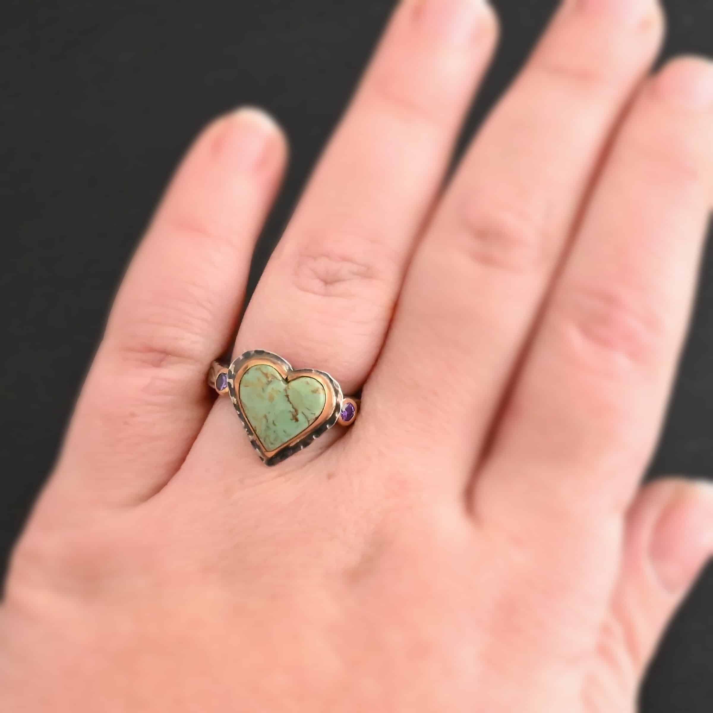 Close up of the "Wild Beauty" Ring. Made out of sterling silver, gold, turquoise, and amethyst. The main stone is a heart shaped turquoise that is set in gold. The turquoise is a mint green color, with lines of gold and brown flowing throughout it. There are two small circular amethysts that are set in gold on the left and right side of the ring. The sterling silver band is engraved with leaves. Ring is worn by a model to demonstrate how large the turquoise is.